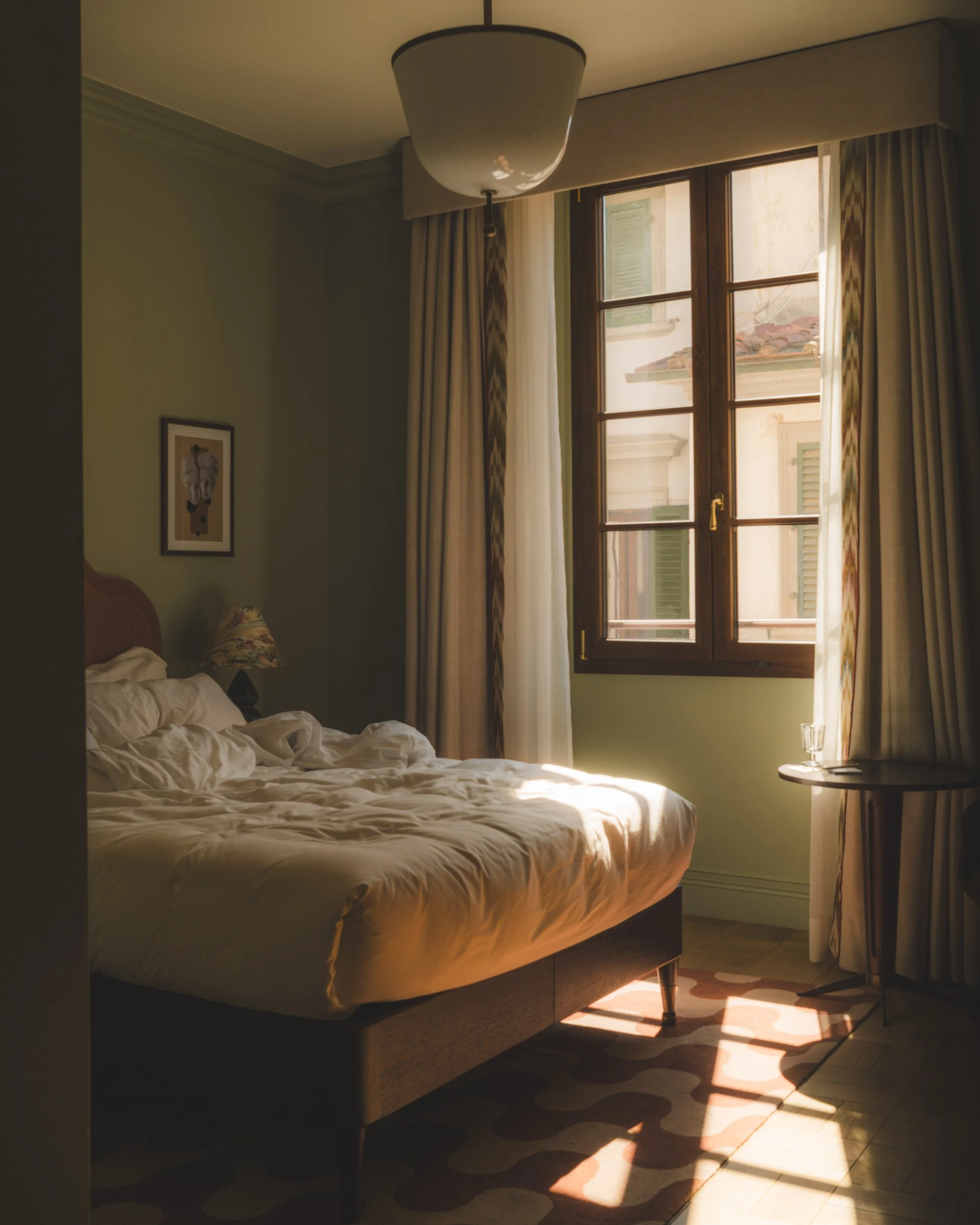 Sunlight coming through a large window with wooden frames, curtains, and shutters, illuminating a bedroom with an unmade bed, a small round side table with a glass of water, a framed picture on the wall, and a patterned rug on the floor.