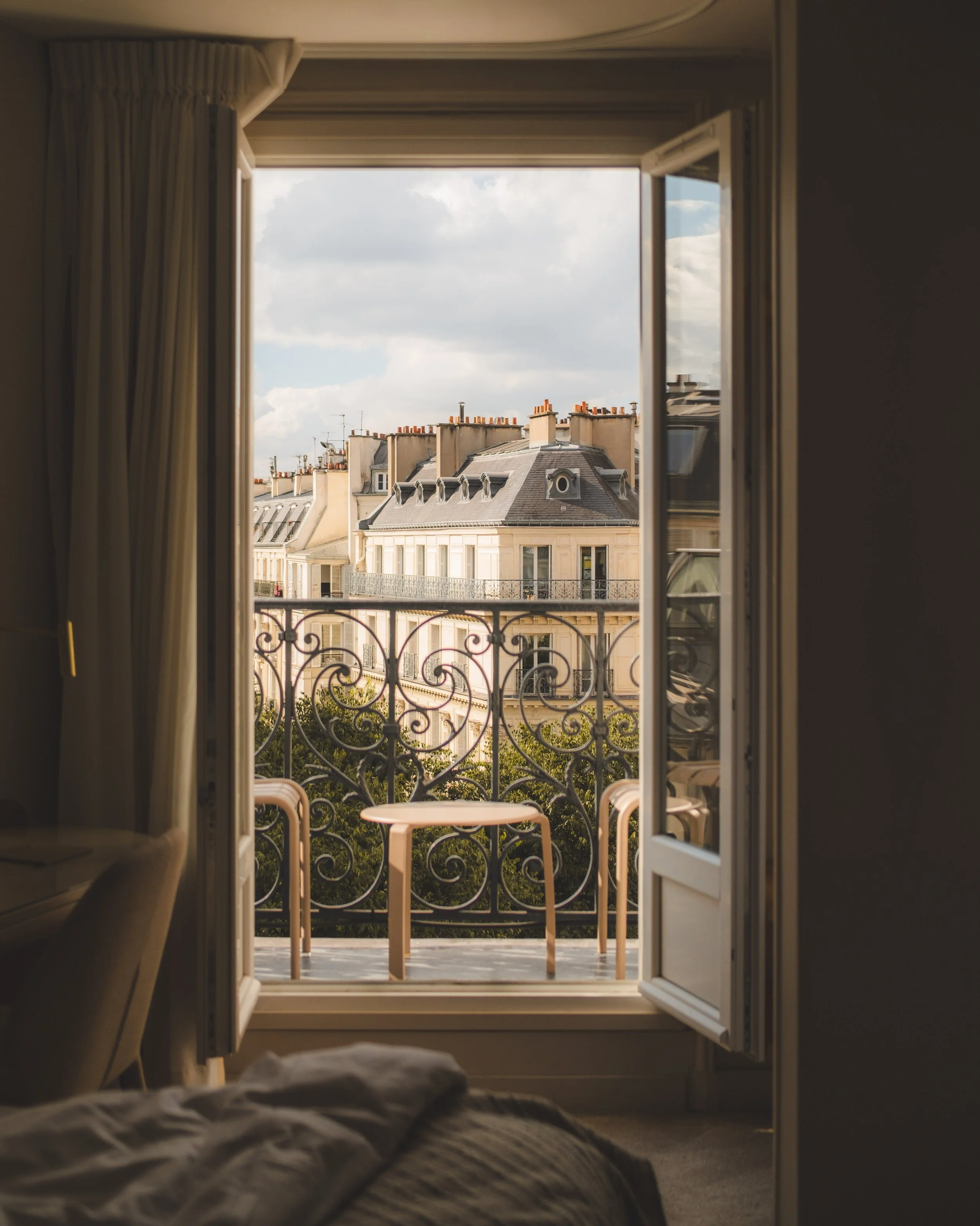 View from a bedroom looking through open French doors onto a small balcony with two pink chairs and a round table, overlooking neighboring Parisian buildings with grey rooftops and chimneys, under a partly cloudy sky.