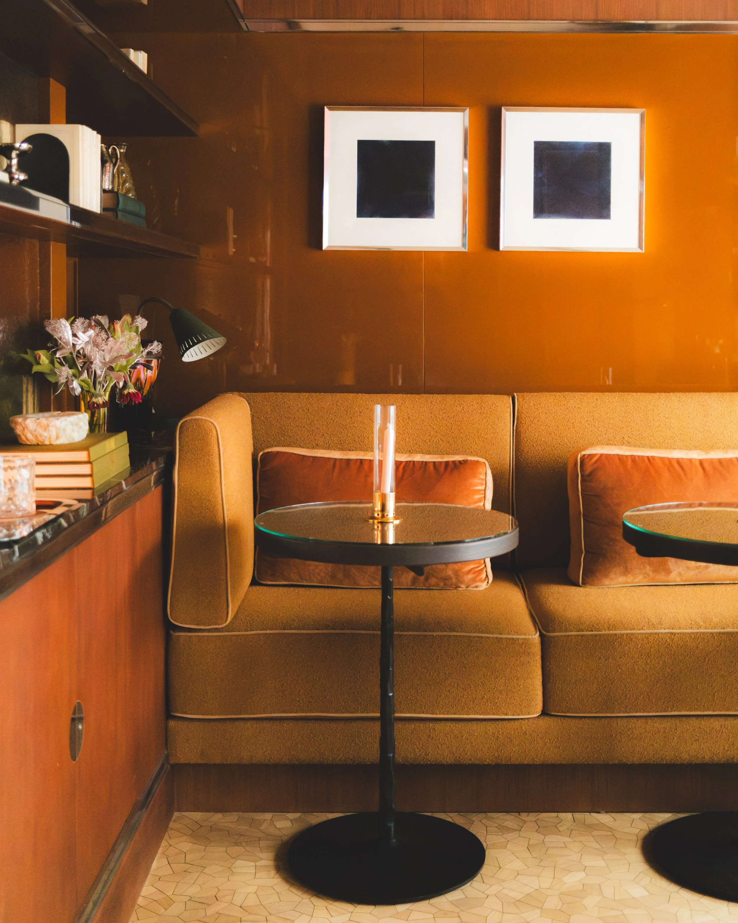 An indoor scene with two framed abstract black and white artworks on a warm orange wall, a round table with a black base and a candle, orange upholstered chairs, a wooden shelf with books and decorations, a floral arrangement, a table lamp, and a tiled floor.