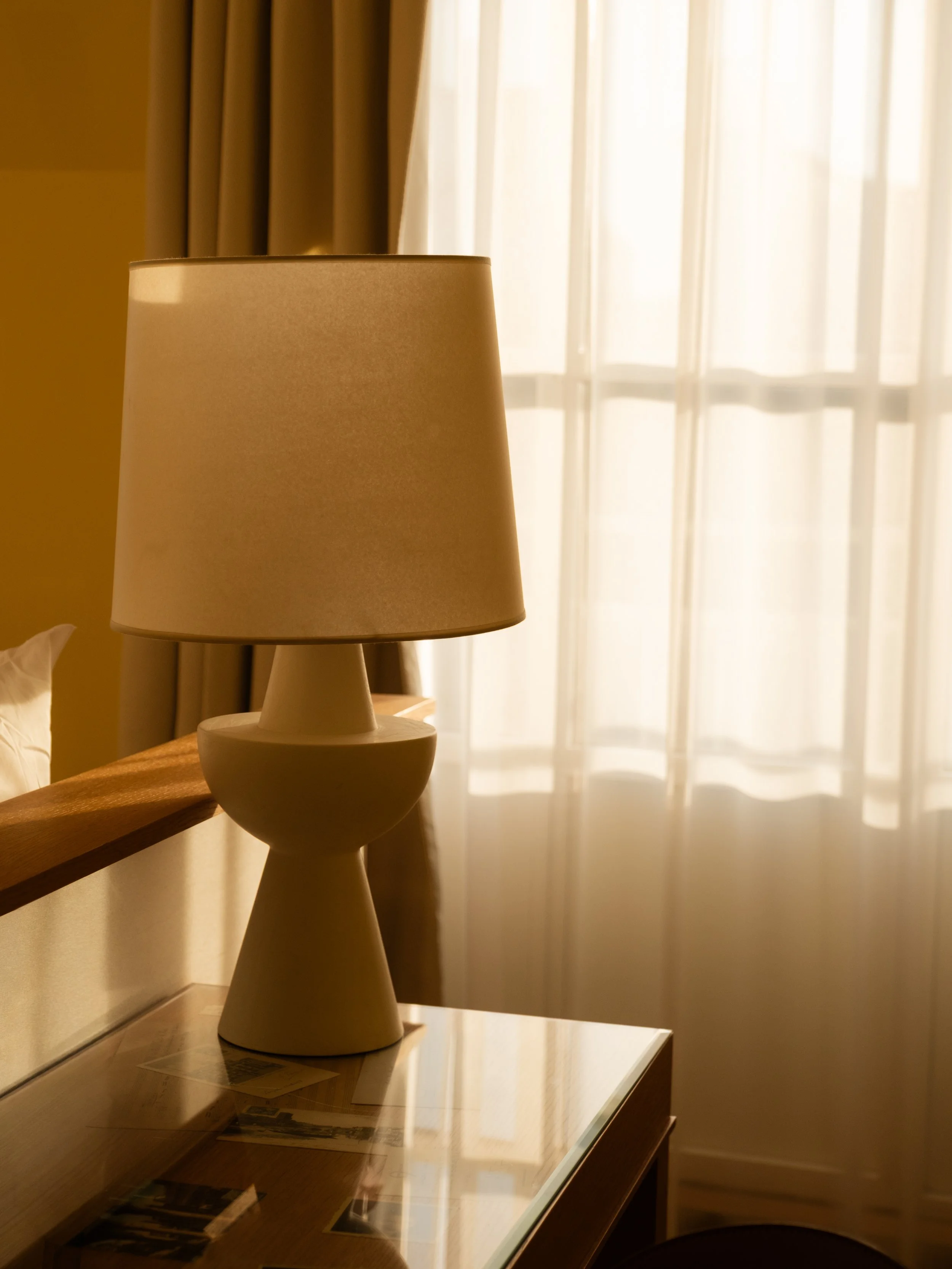A beige table lamp on a wooden nightstand next to a bed, beside a window with sheer curtains, in a softly lit room. Lionel Chu - Hotel & Travel Photography