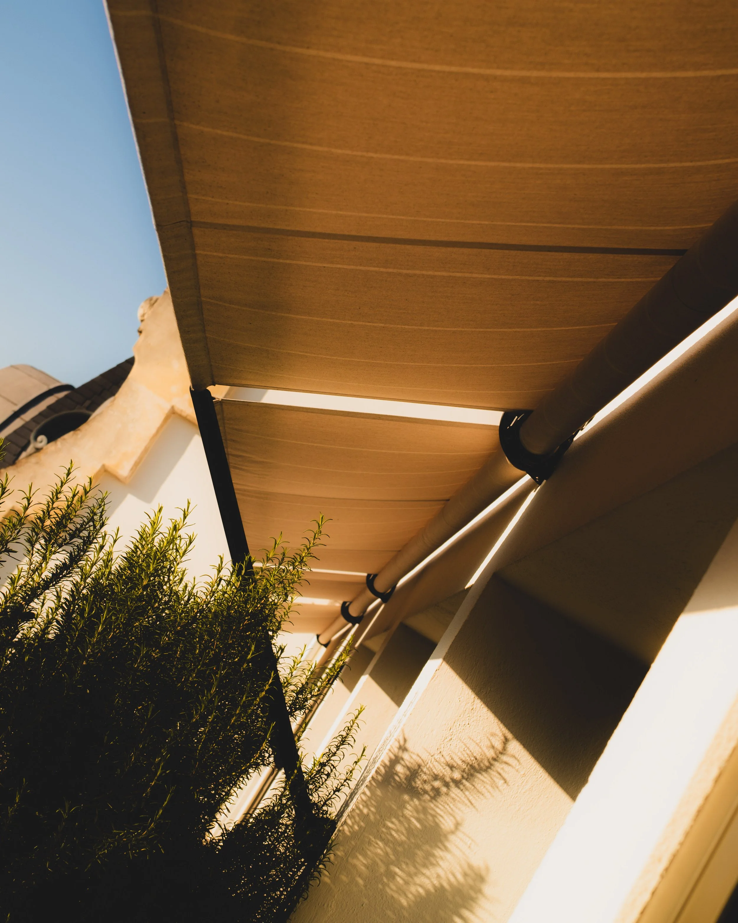 A view of a wooden roof or overhang, cast shadows on a beige wall, with some greenery and a small chimney or vent in the background, during sunset or late afternoon.