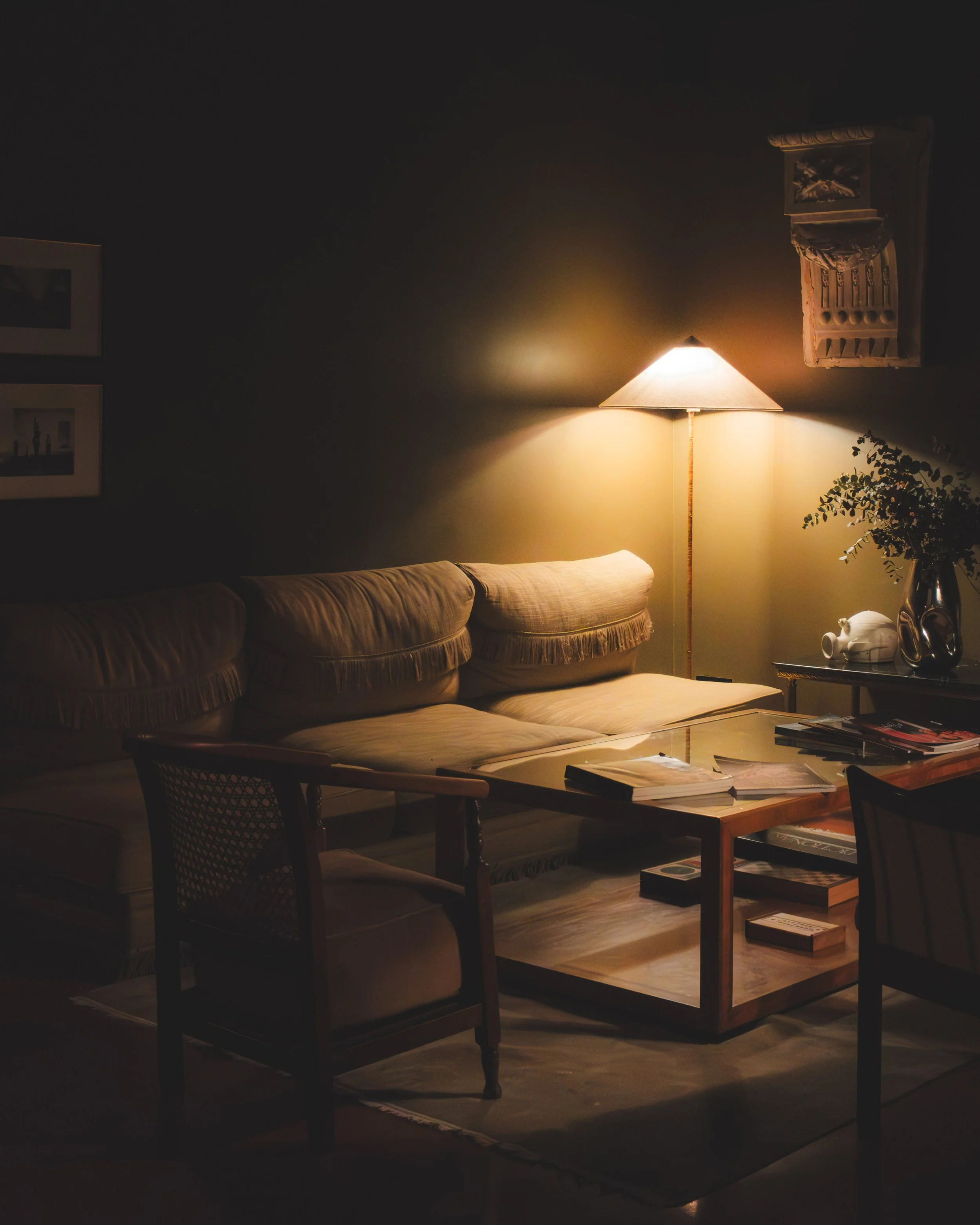 Hotel lobby with beige sofa, wooden coffee table, table lamp, wall-mounted shelf, potted plant, and pictures on wall, dimly lit.