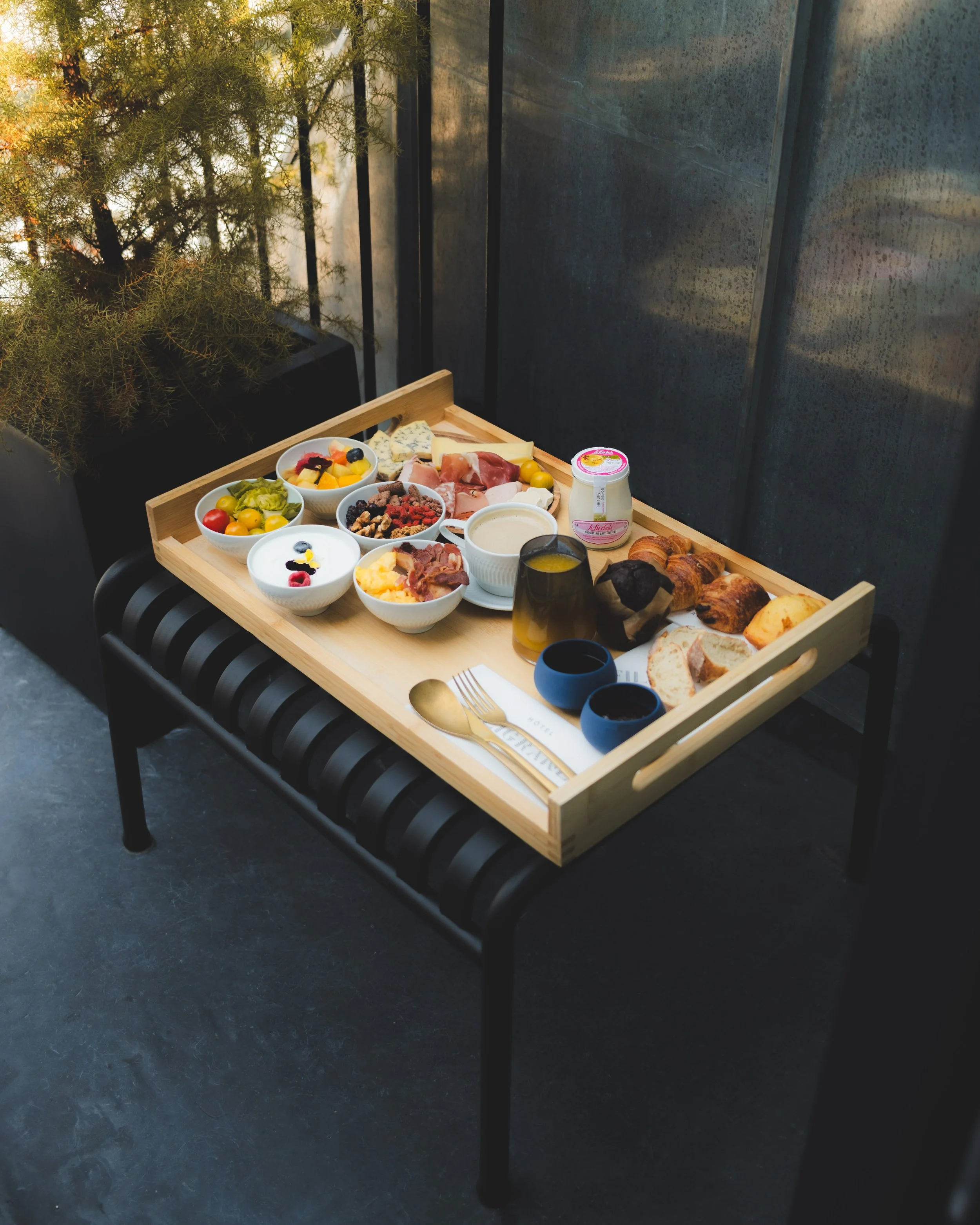 A tray with various breakfast items including bowls of yogurt with fruit, cheeses, cold cuts, croissants, bread, and a jar of yogurt, set on a black bench outside with a potted plant and gray wall in the background.