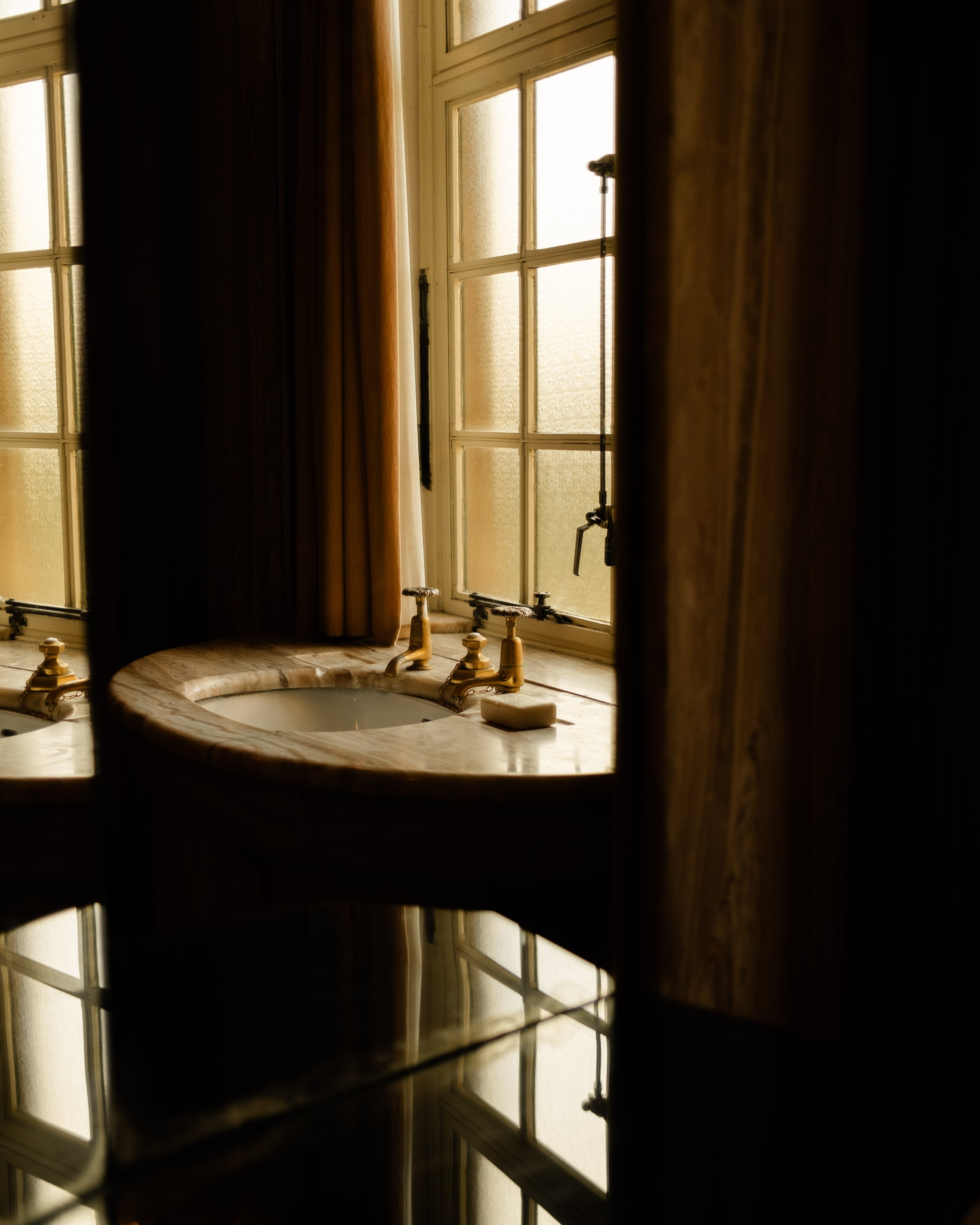 A vintage bathroom with a marble countertop, gold faucet fixtures, and a large window with frosted glass and curtains, seen through a partially opened door. Lionel Chu - Hotel & Travel Photographer