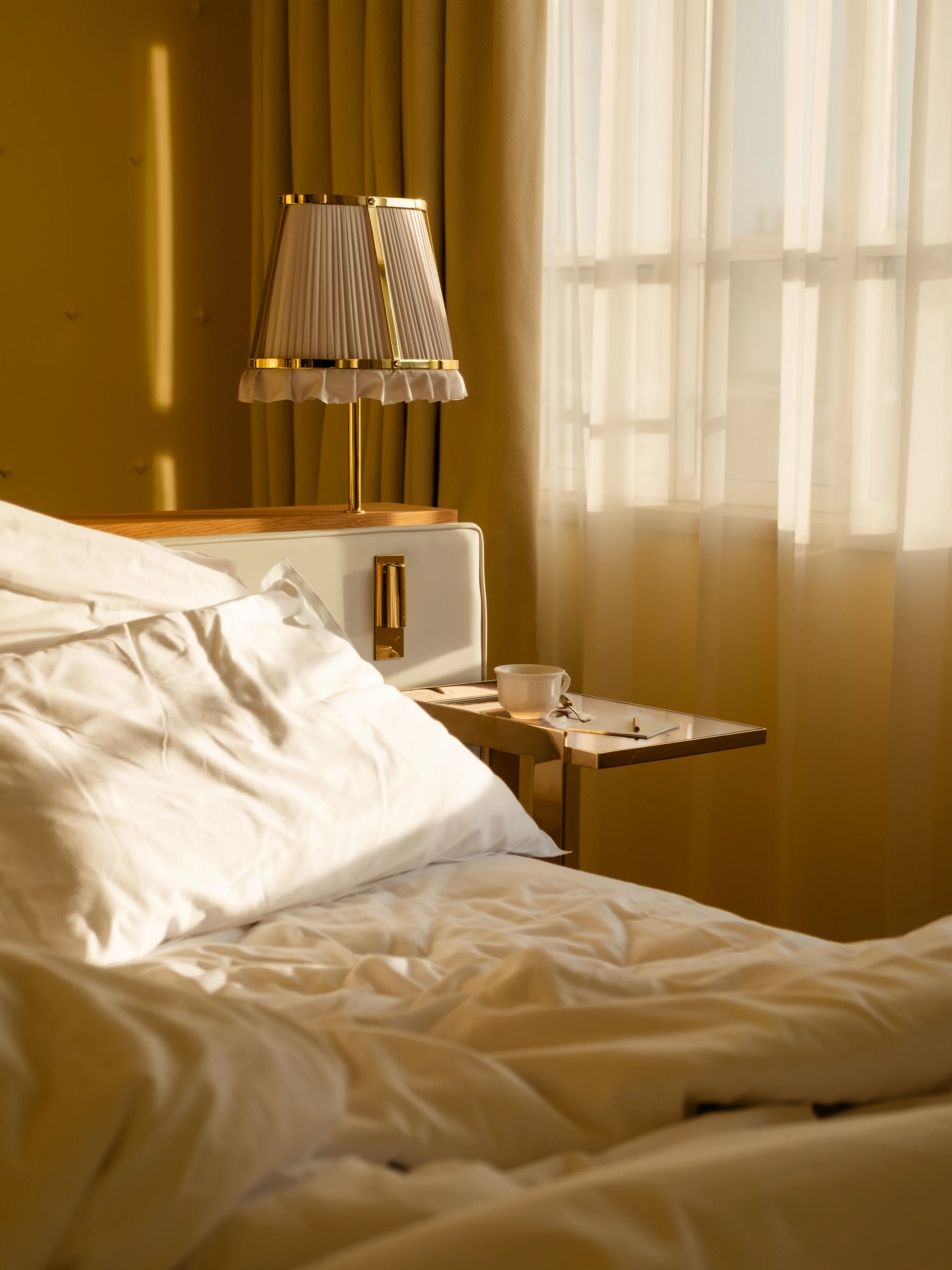 A bedroom with white bed linens, a nightstand, a wall-mounted lamp with a pleated lampshade, and cream-colored curtains letting in soft natural light.