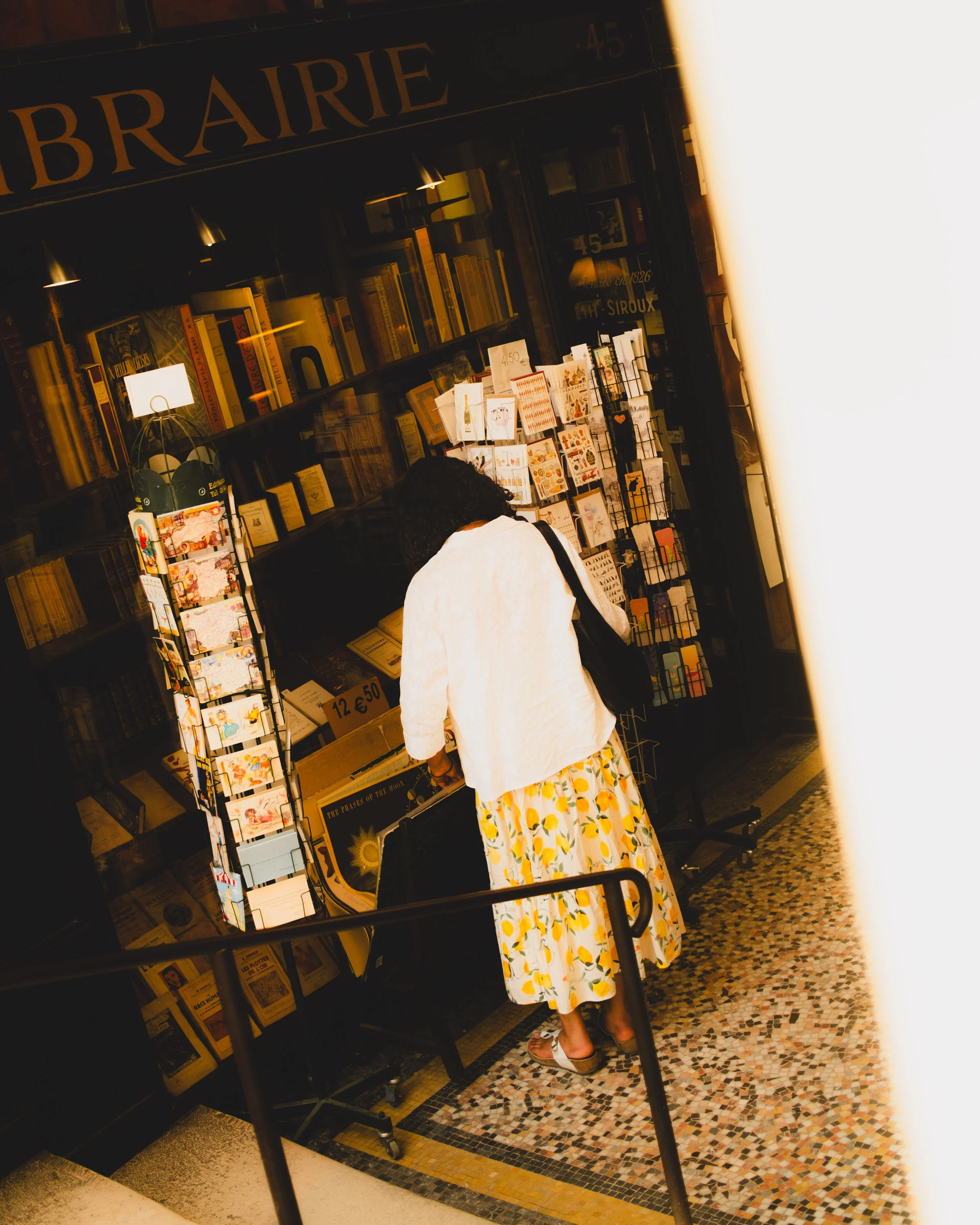A woman shopping at a bookstore, standing in front of a display of postcards and books, wearing a white jacket and a floral skirt. Lionel Chu - Hotel & Travel Photographer
