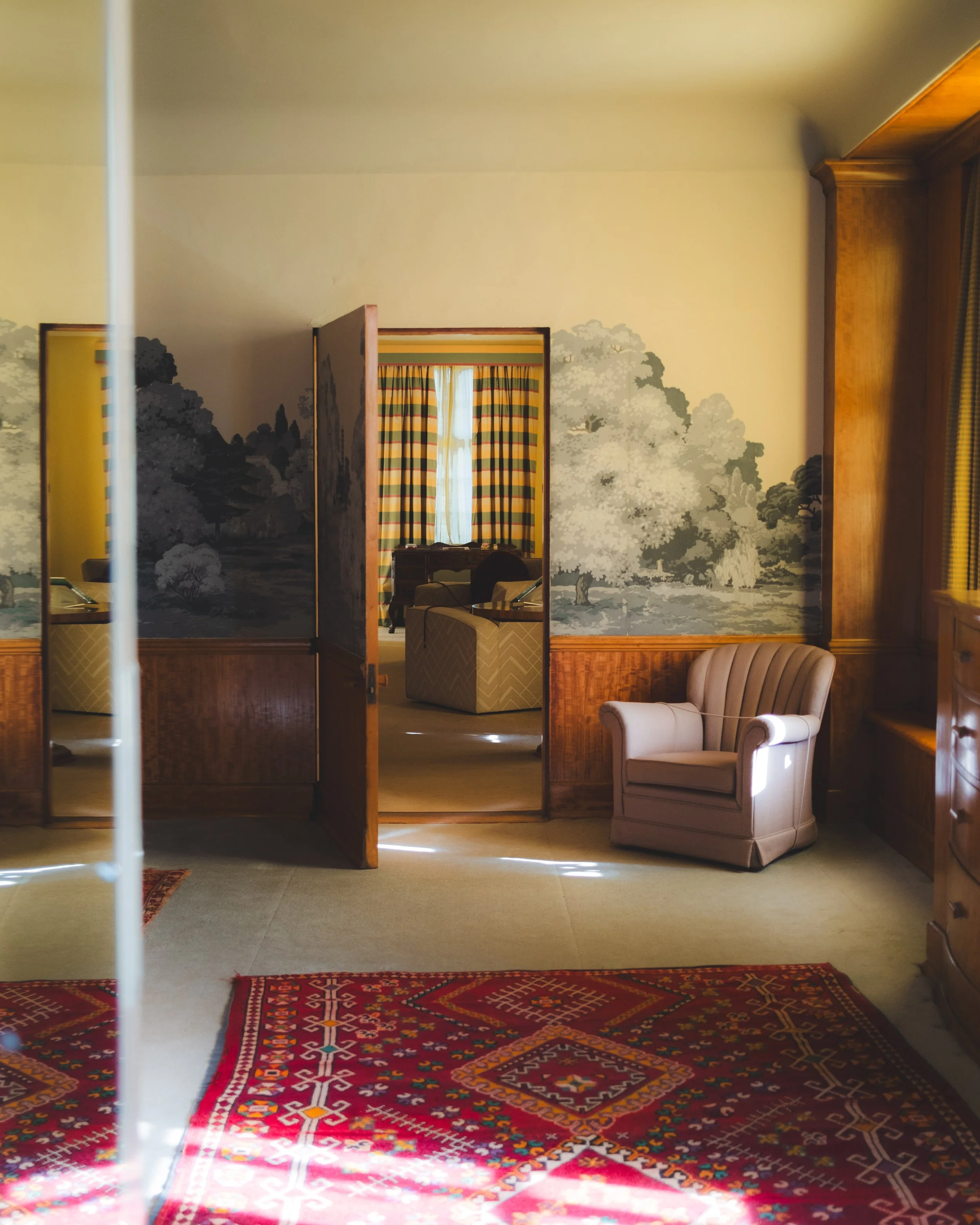 An interior room with a red patterned rug on the floor, a beige armchair, and a wall decorated with a mural of trees and landscape. There is a doorway with striped curtains leading to another room, and a large wooden frame on the wall.