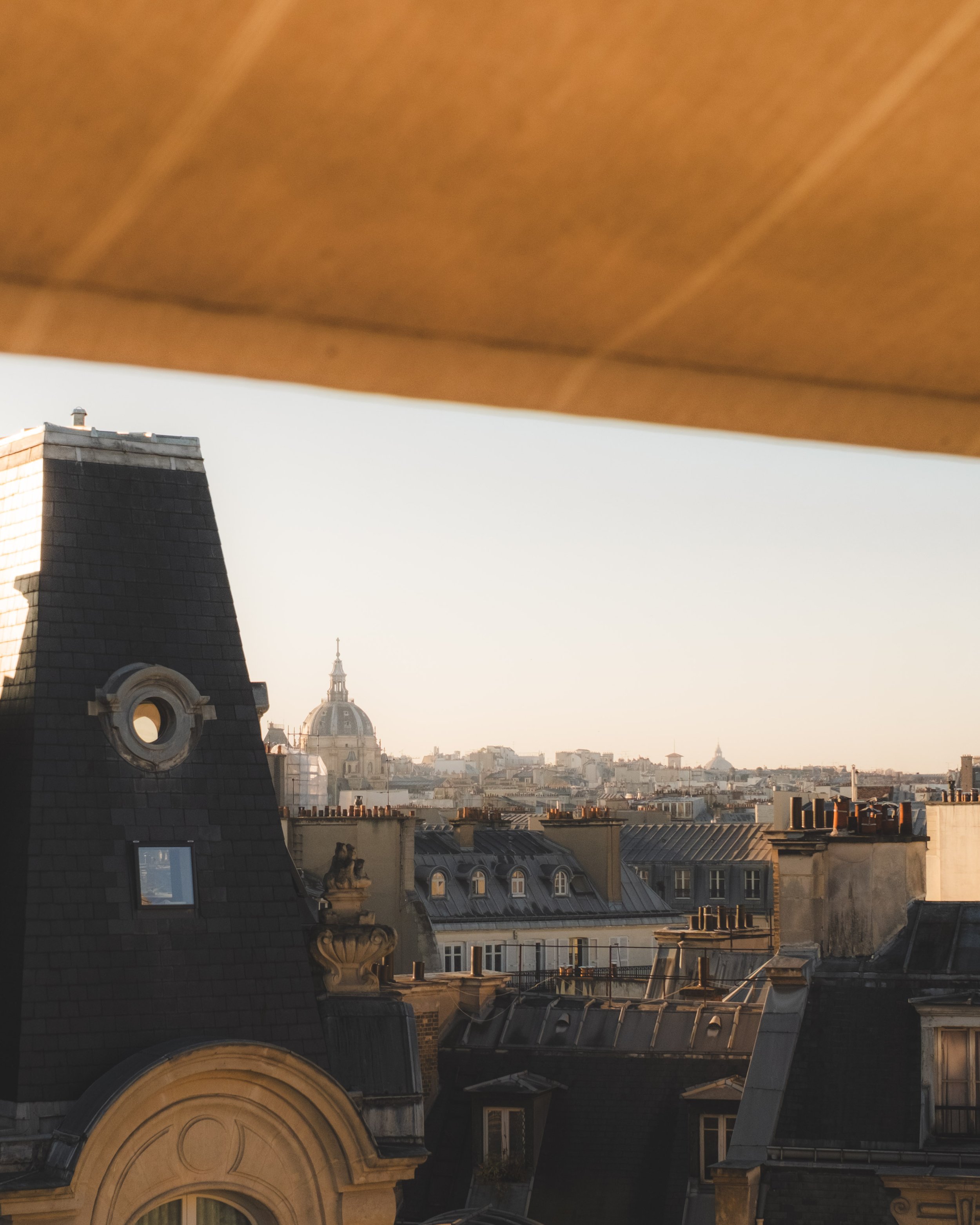 Paris cityscape with rooftop buildings, a church dome in the distance, and a rooftop chimney, under a partially visible wooden ceiling.