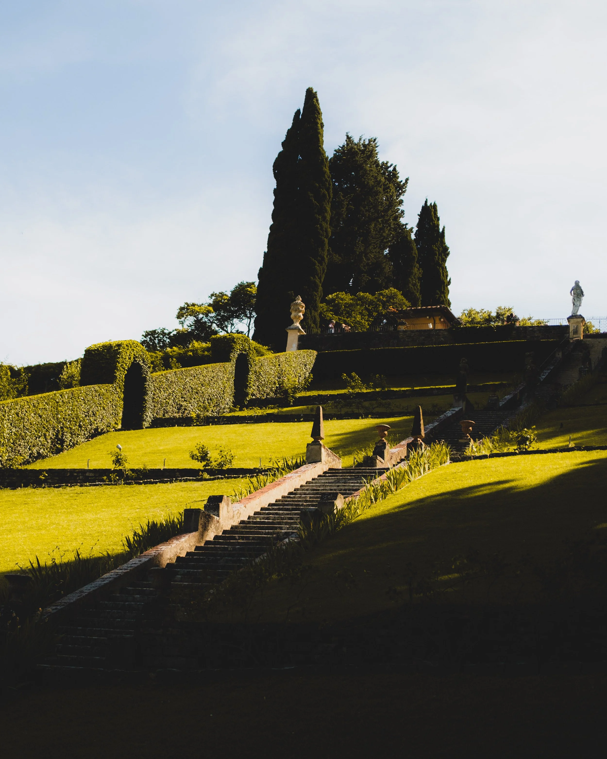 A series of stairs ascend a lush, terraced hillside garden with tall cypress trees and statues, under a cloudy sky. Lionel Chu - Hotel & Travel Photographer