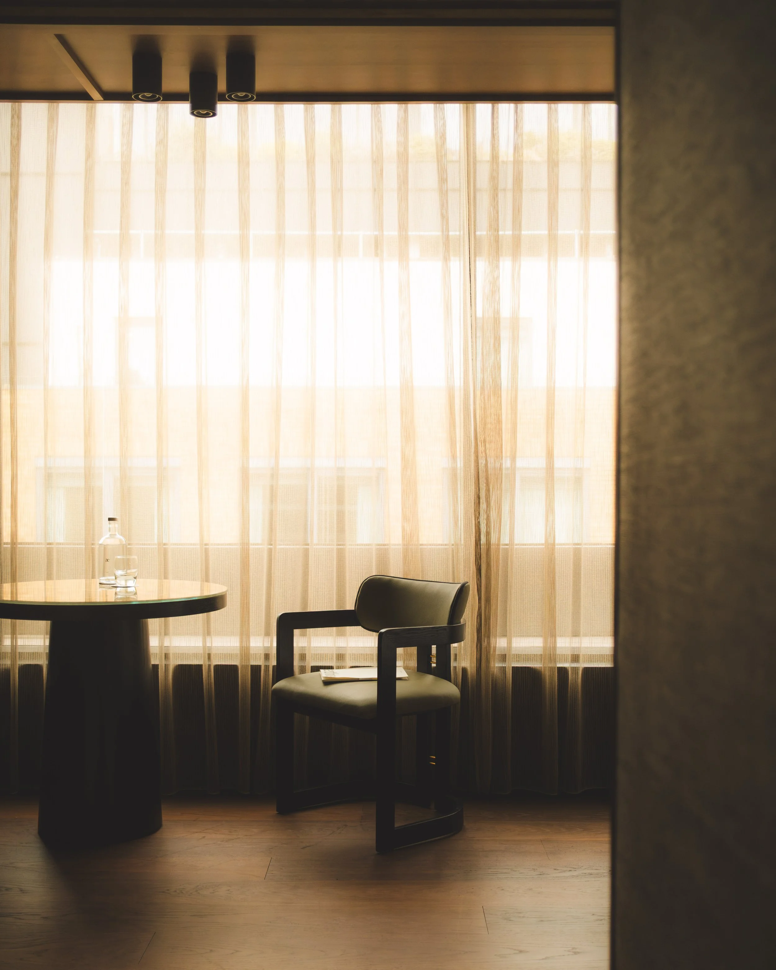 A round table with a water bottle and glass placed on top, and an empty chair in front of a large window with sheer beige curtains. Sunlight filters through the curtains, illuminating the room.