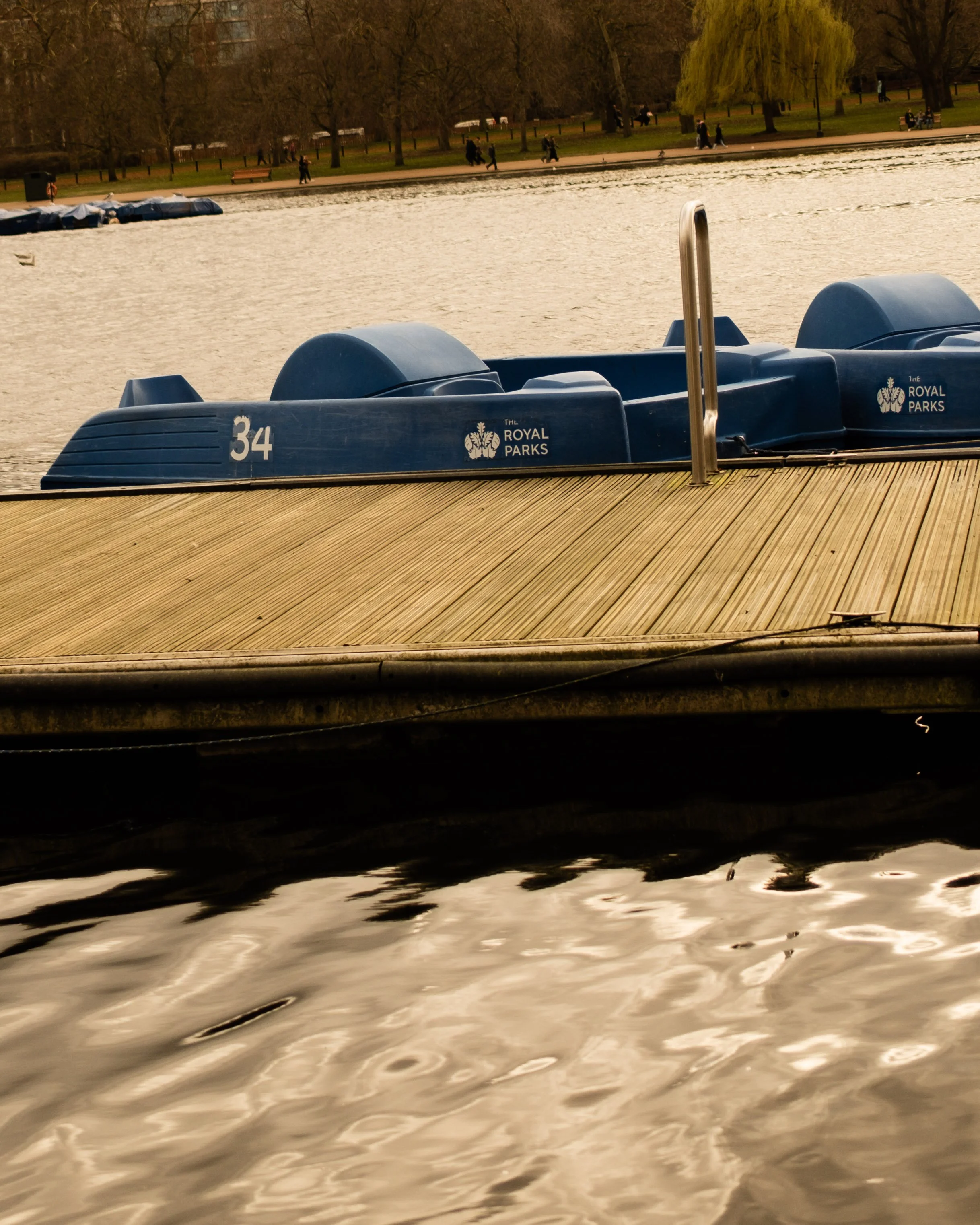 A blue paddle boat docked at a wooden pier on a calm lake in a park, with trees and walking paths visible in the background. Lionel Chu - Hotel & Travel Photographer