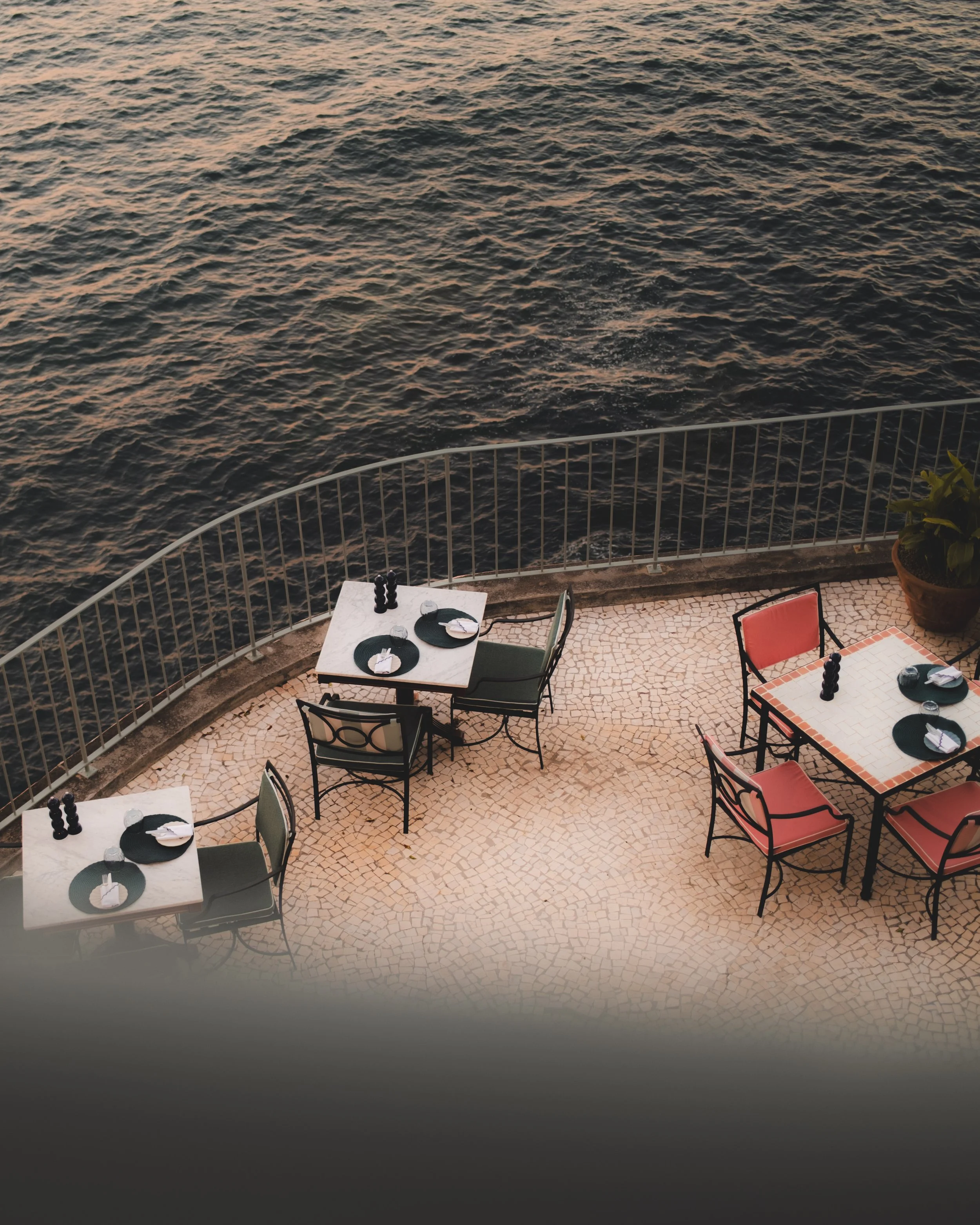Outdoor café on a terrace overlooking water, with several tables and chairs set for dining, and a railing along the edge.