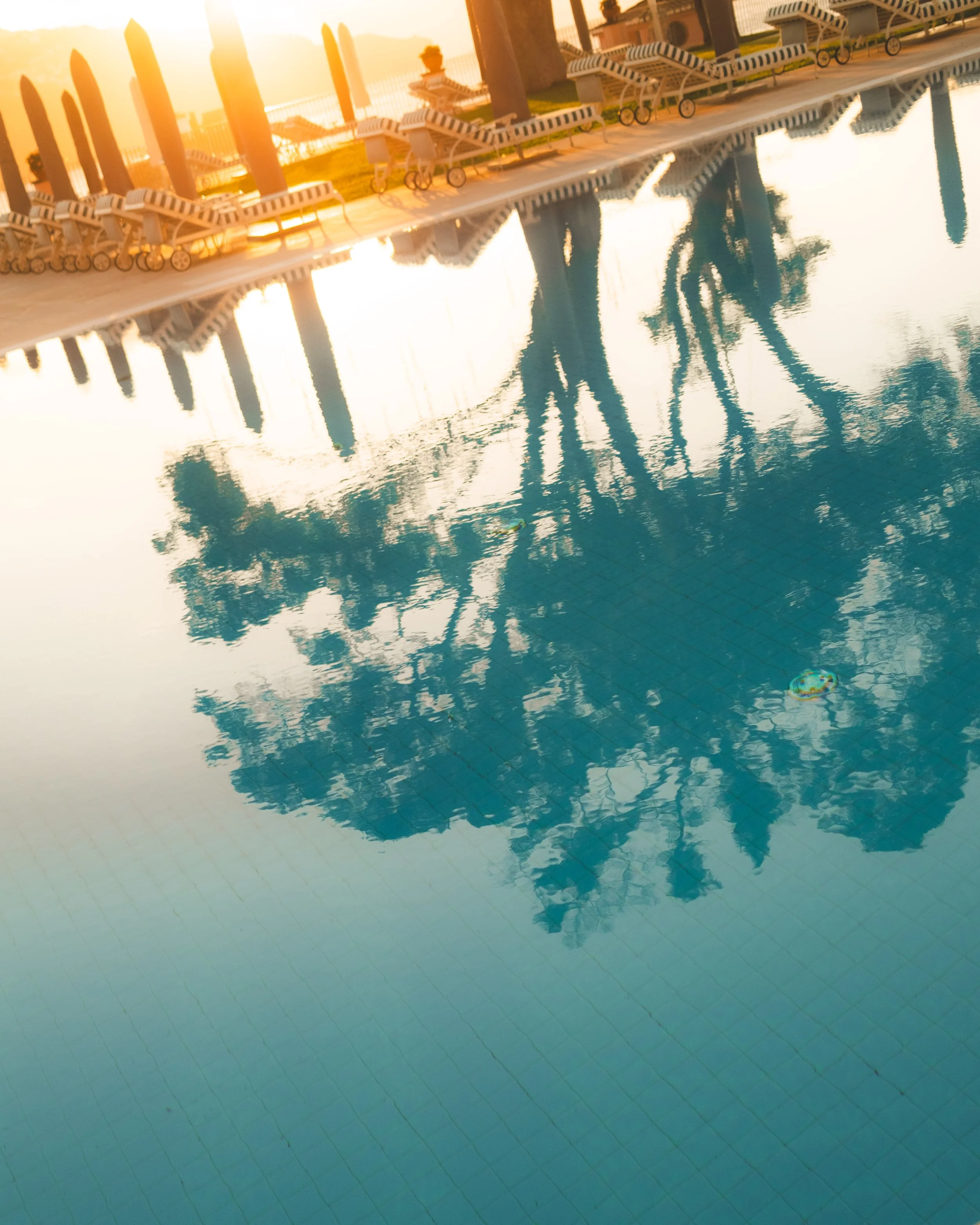 A swimming pool with lounge chairs along the edge, reflecting the sky and nearby structures, during sunset.