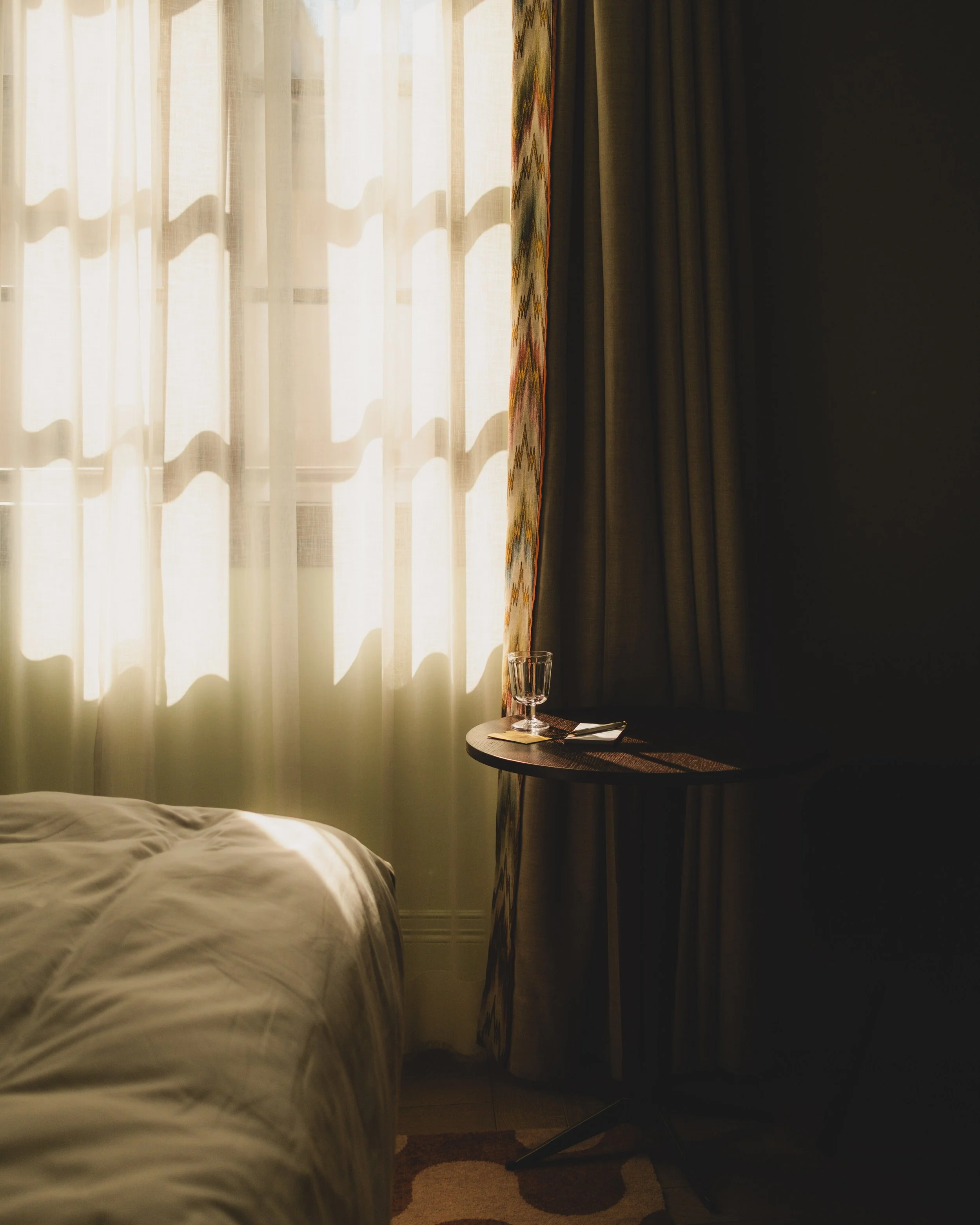 Sunlight streams through white curtains in a hotel room, casting shadows. There is a small round table with a glass of water, a napkin, and a fork. The bed with white linens is partially visible in the foreground, and dark curtains are partially drawn on the right side.