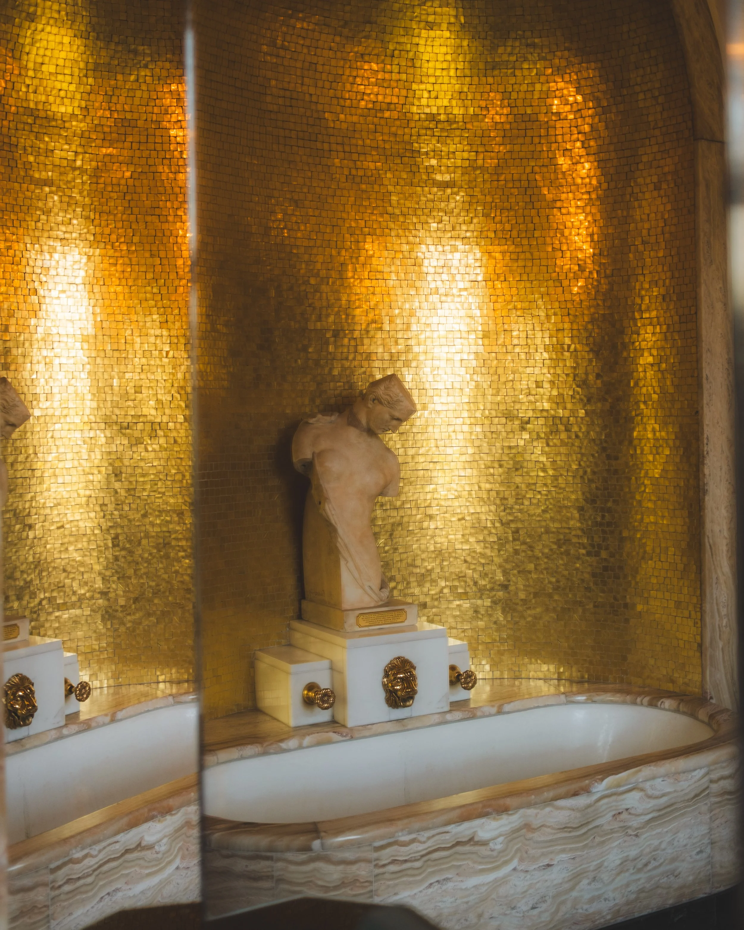 A marble statue of a headless man on a rectangular pedestal in front of a golden mosaic wall with ornate gold lion head fixtures.