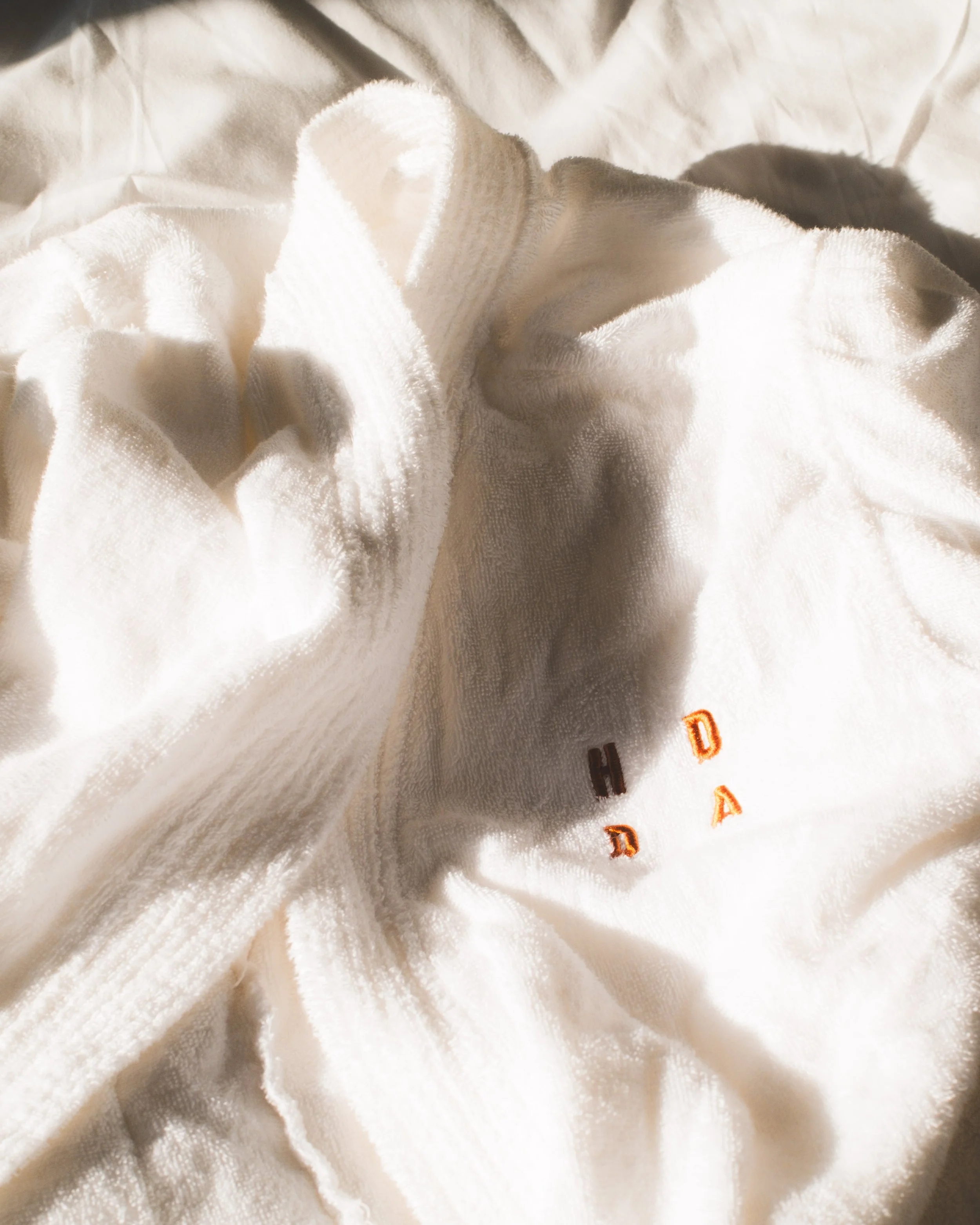 Close-up of an hotel dressing gown with embroidered orange text, placed on a soft, crumpled white fabric background.