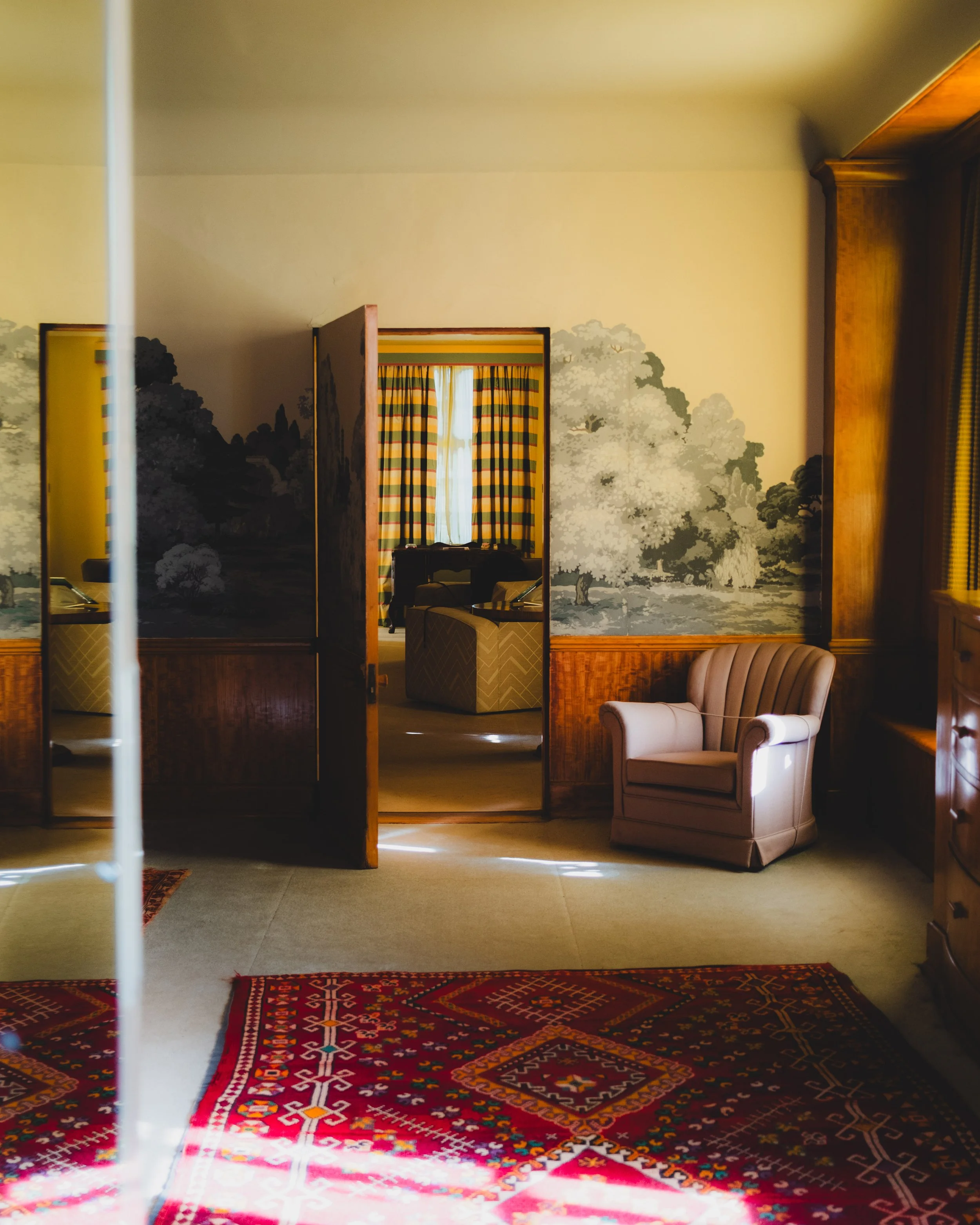 An interior view of a vintage manor bedroom with a red patterned rug, beige armchair, and decorated wallpaper featuring trees and landscape scene, seen through an open door. Lionel Chu - Hotel & Travel Photographer