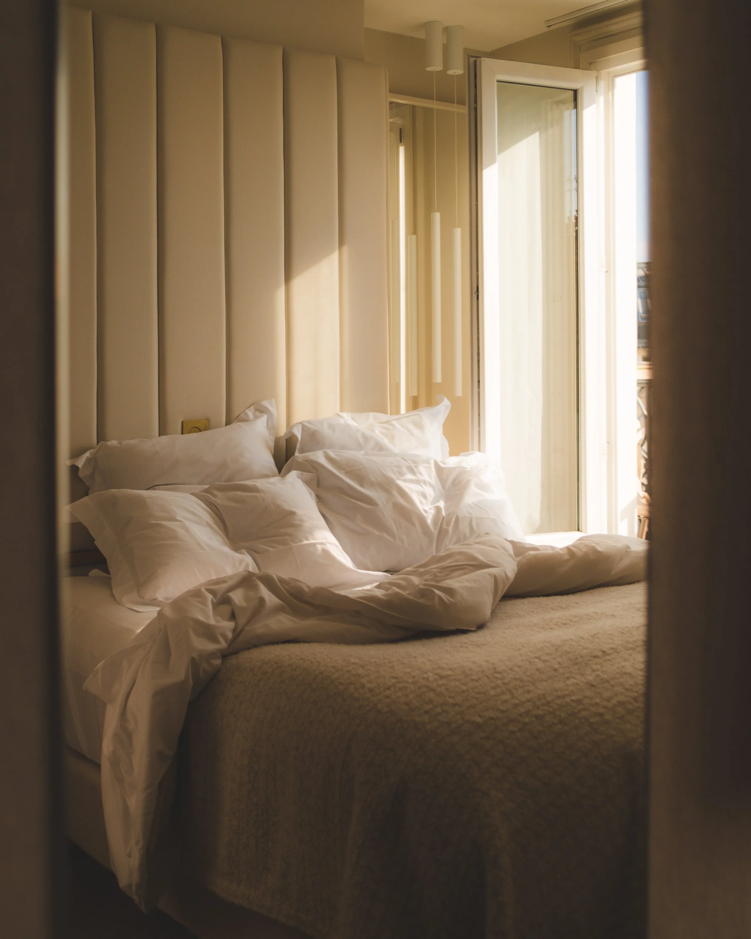 A disorganized bed with white bedding, partially pushed aside, sitting on a beige carpet in a sunlit room with large windows and a cream-colored padded headboard.