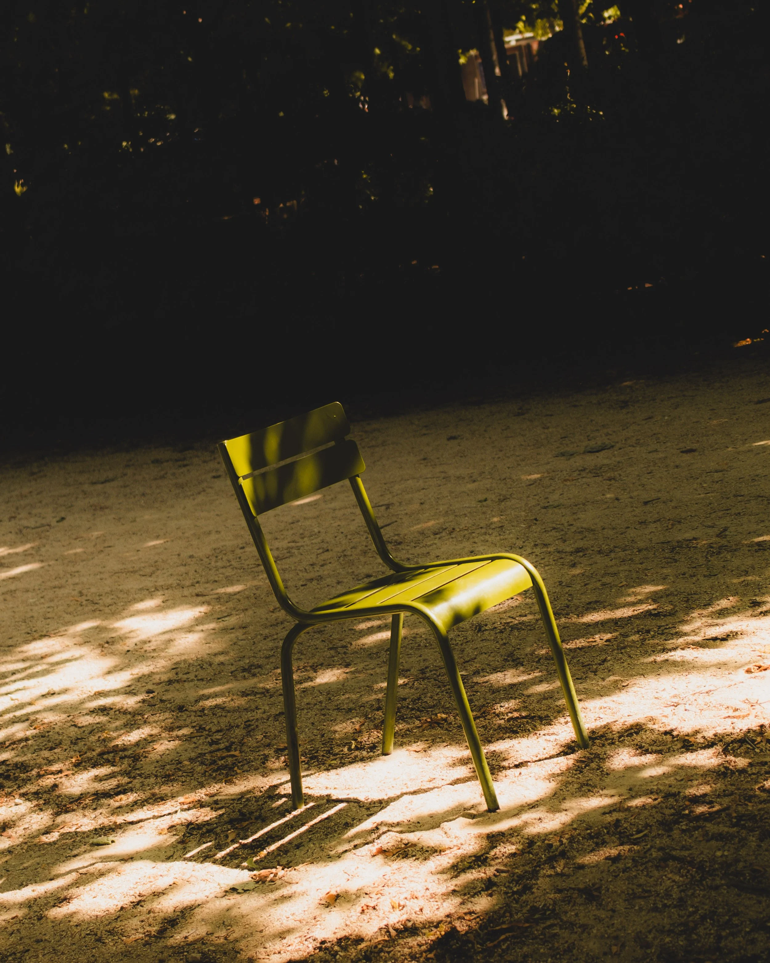 An empty green metal chair on a dirt ground, casting shadows, in a shaded outdoor area surrounded by trees. Lionel Chu - Hotel & Travel Photographer
