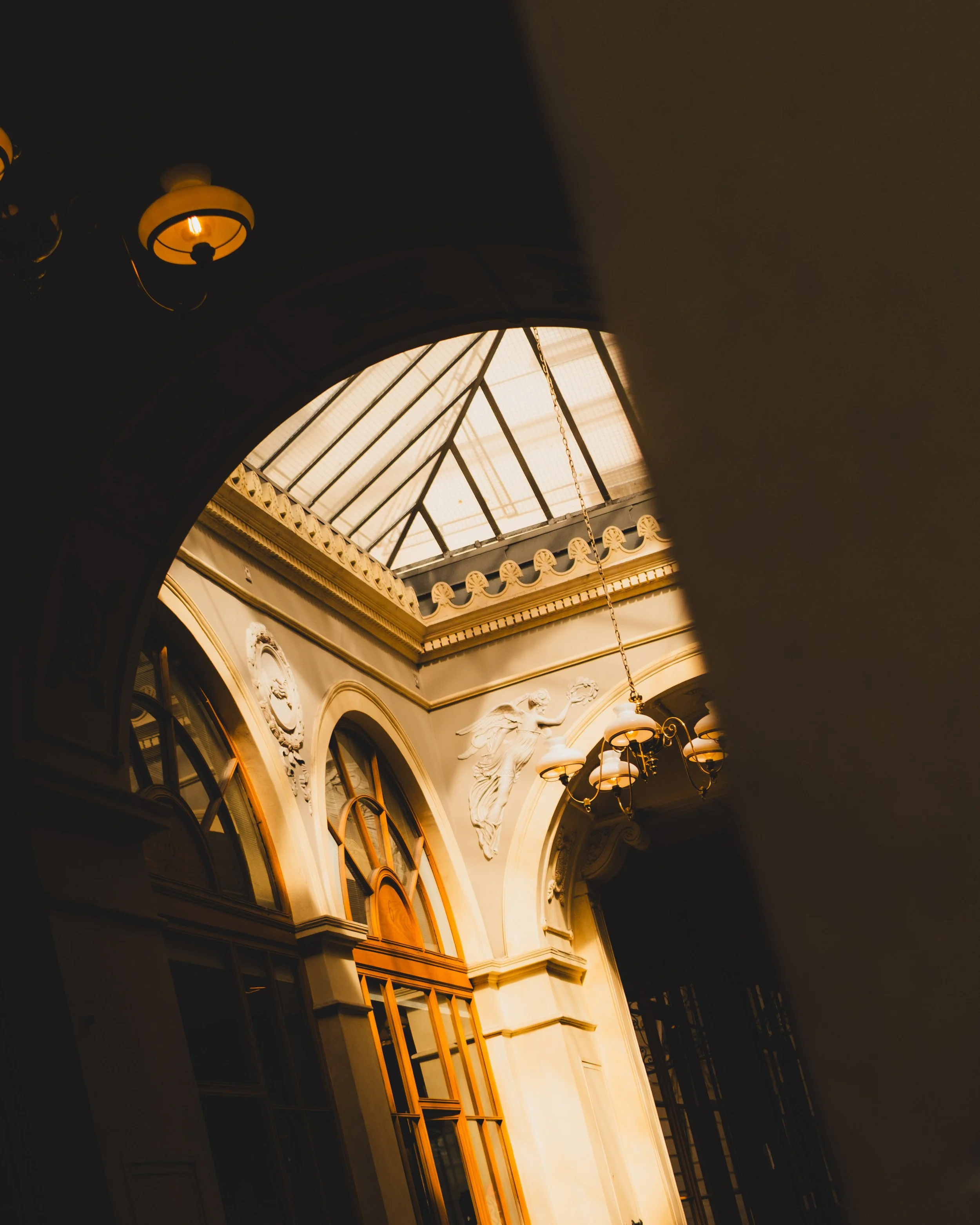 Interior view of an ornate building with large arched windows, decorative ceiling, and a statue of a woman with flowing drapery near the corner, illuminated by natural light from a glass ceiling. Lionel Chu - Hotel & Travel Photographer.