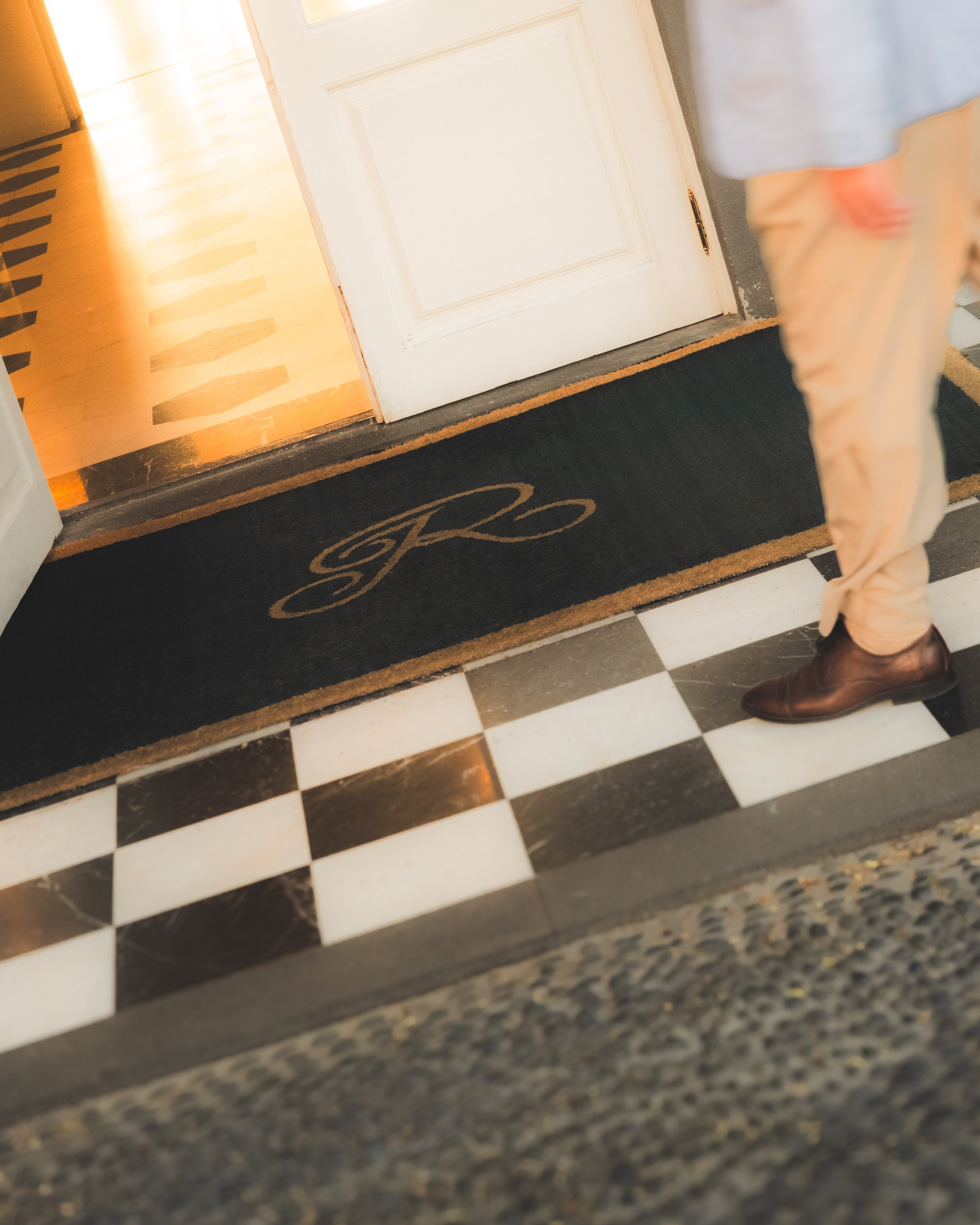 Part of a person's lower body and legs in tan pants and brown shoes standing on a black and white checkered tile floor next to a black mat with a gold monogram and a white door. The door has a gold handle and is partially open, revealing a warm yello