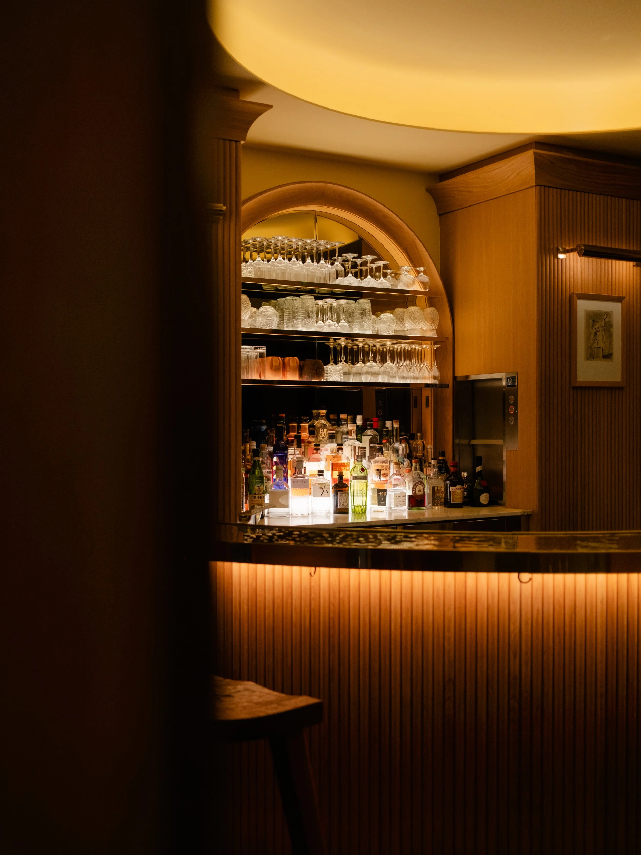 A dimly lit hotel bar with shelves of glasses of various types and bottles of alcohol, featuring warm wood paneling and ambient lighting. Lionel Chu - Hotel & Travel Photography