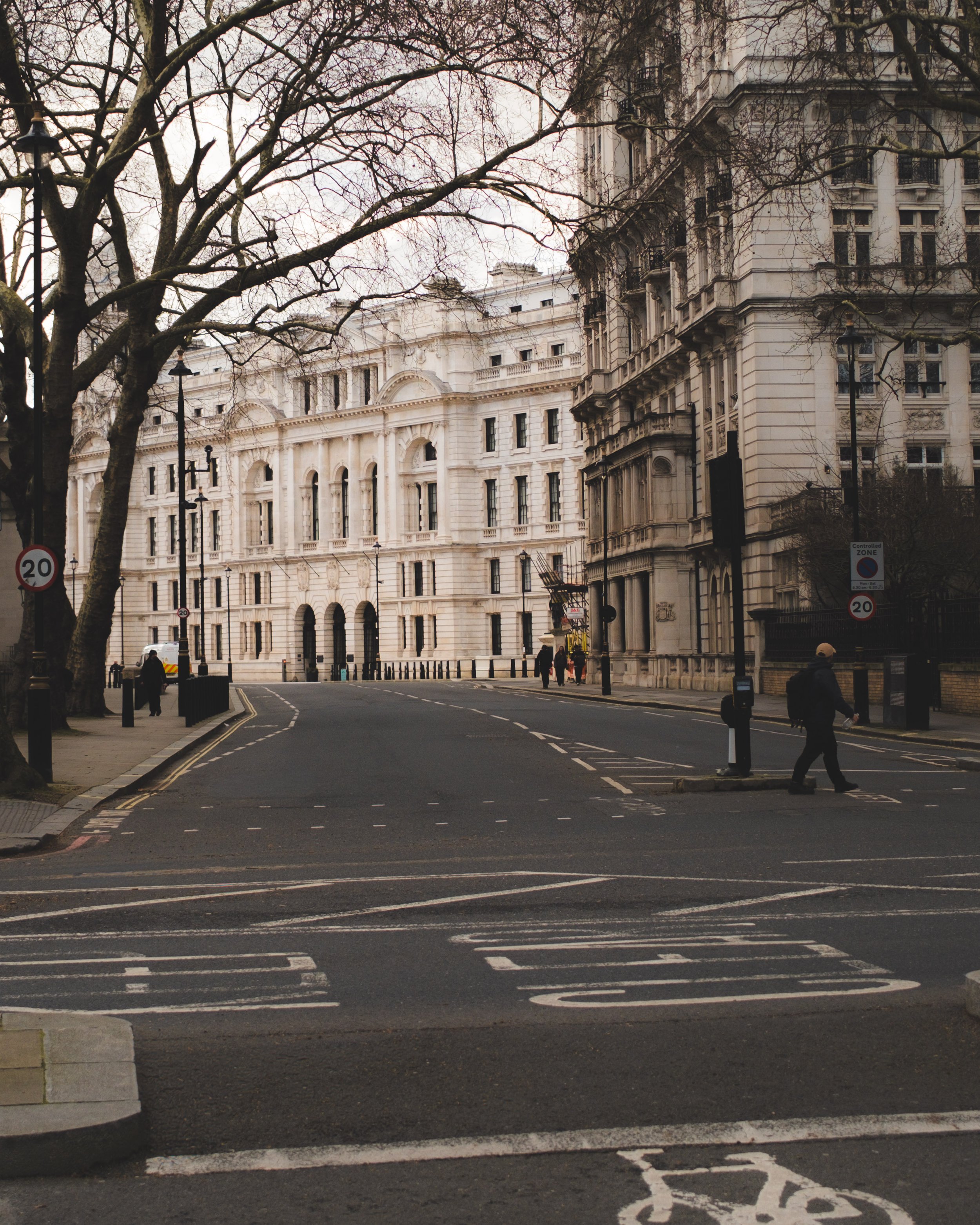 Street view with historical white building, leafless trees, street signs, and pedestrians during cloudy day.
