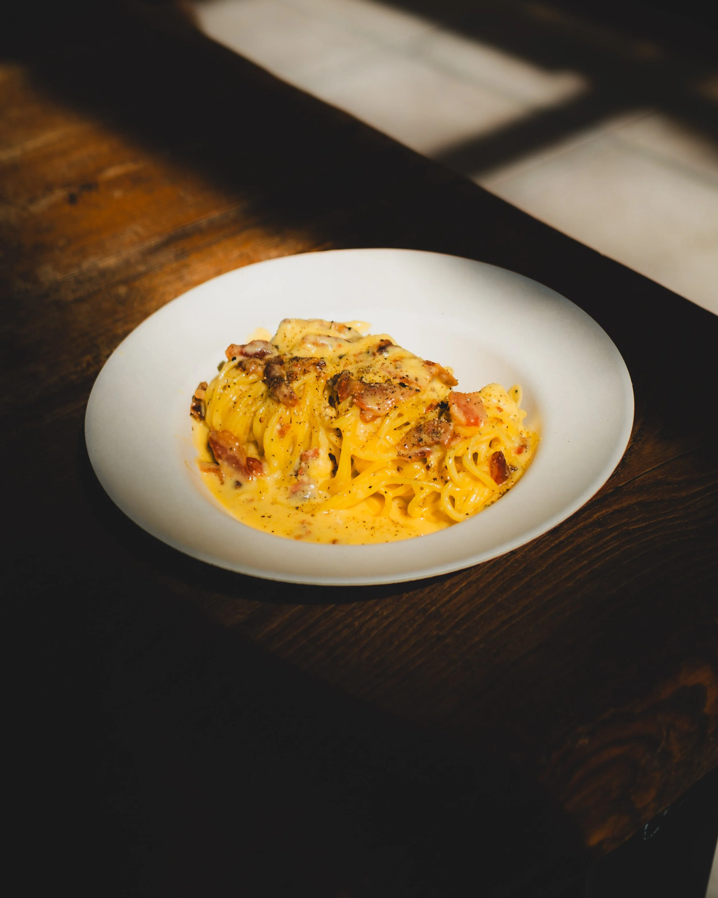 A white bowl of carbonara pasta with guanciale bits on a wooden table. Lionel Chu - Hotel & Travel Photographer