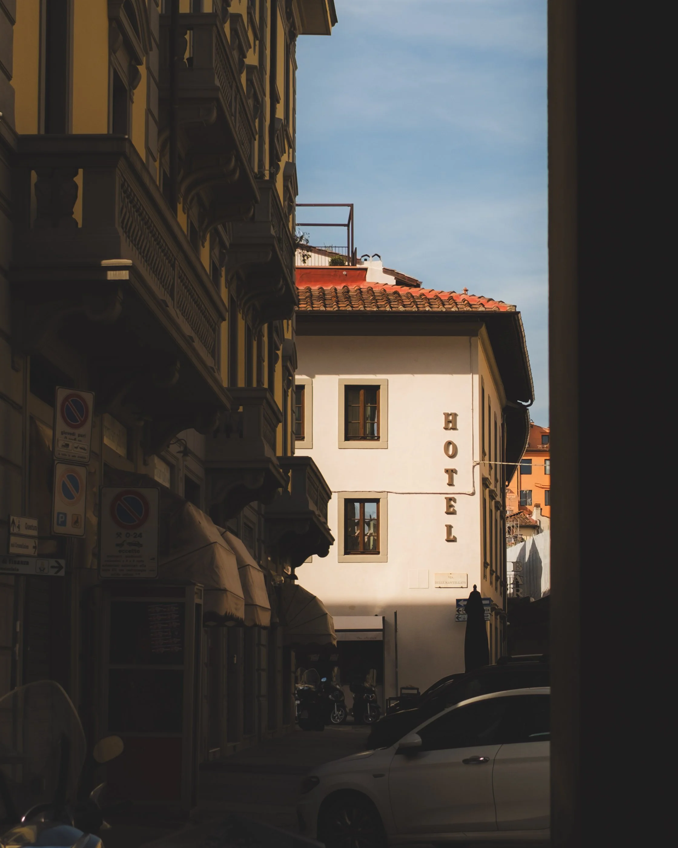 A street view looking past a dark opening, showing buildings with architectural details, a hotel sign, parked cars, and motorcycles in a European city under a blue sky.