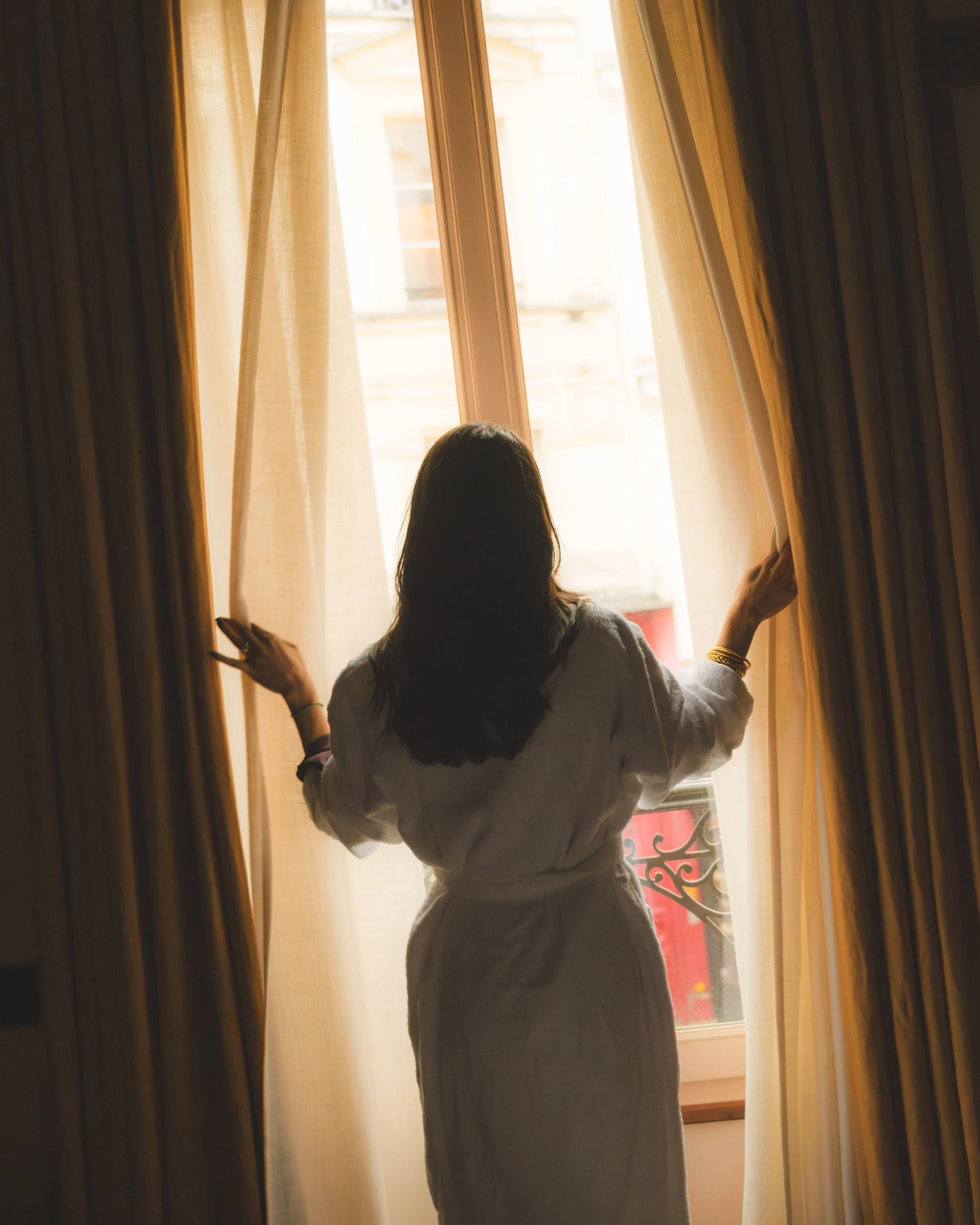Woman standing near a window, partially covered by beige curtains, with sunlight streaming in.
