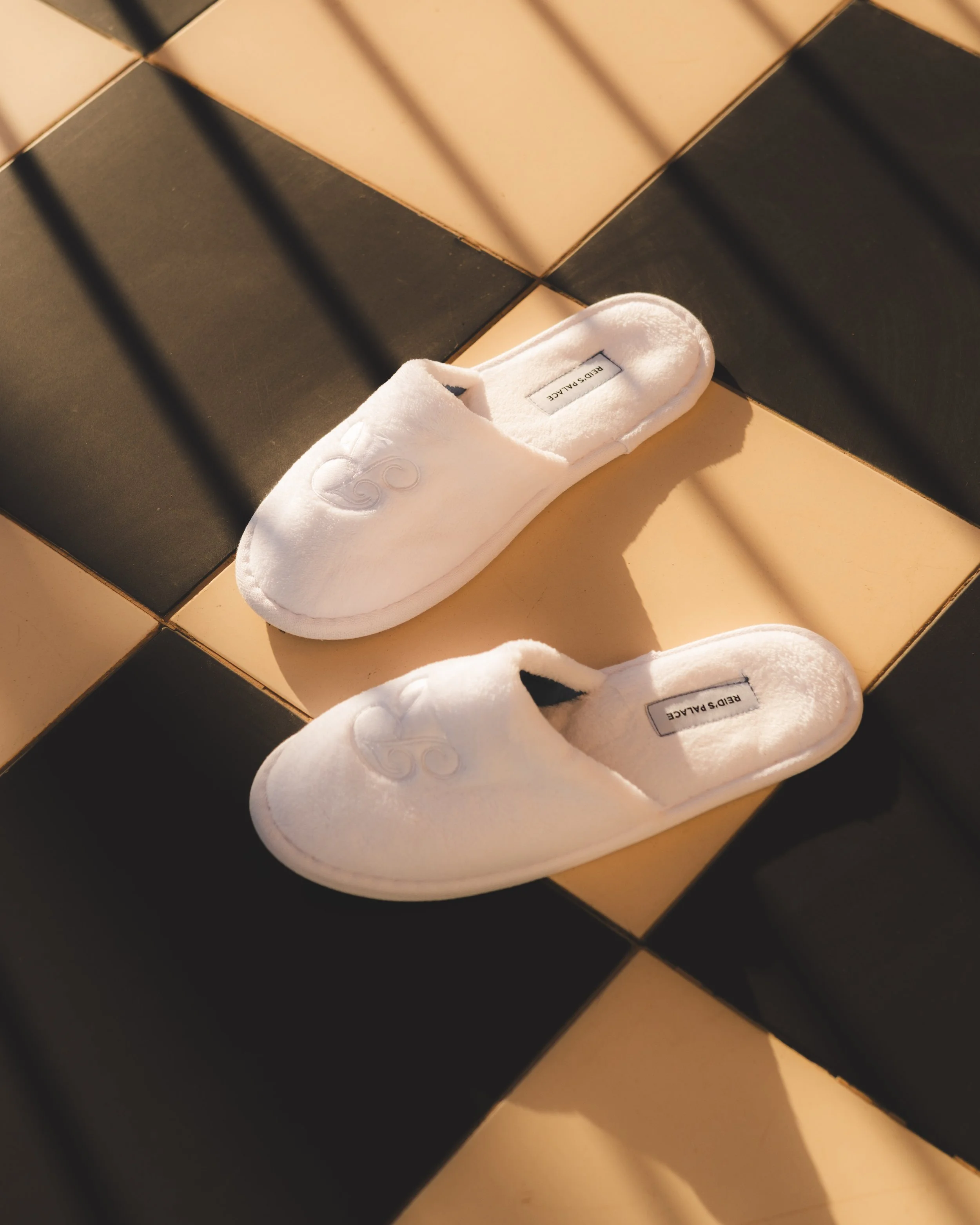 White plush slippers with 'REIDS PLACE' labels sitting on black and beige tiled floor.