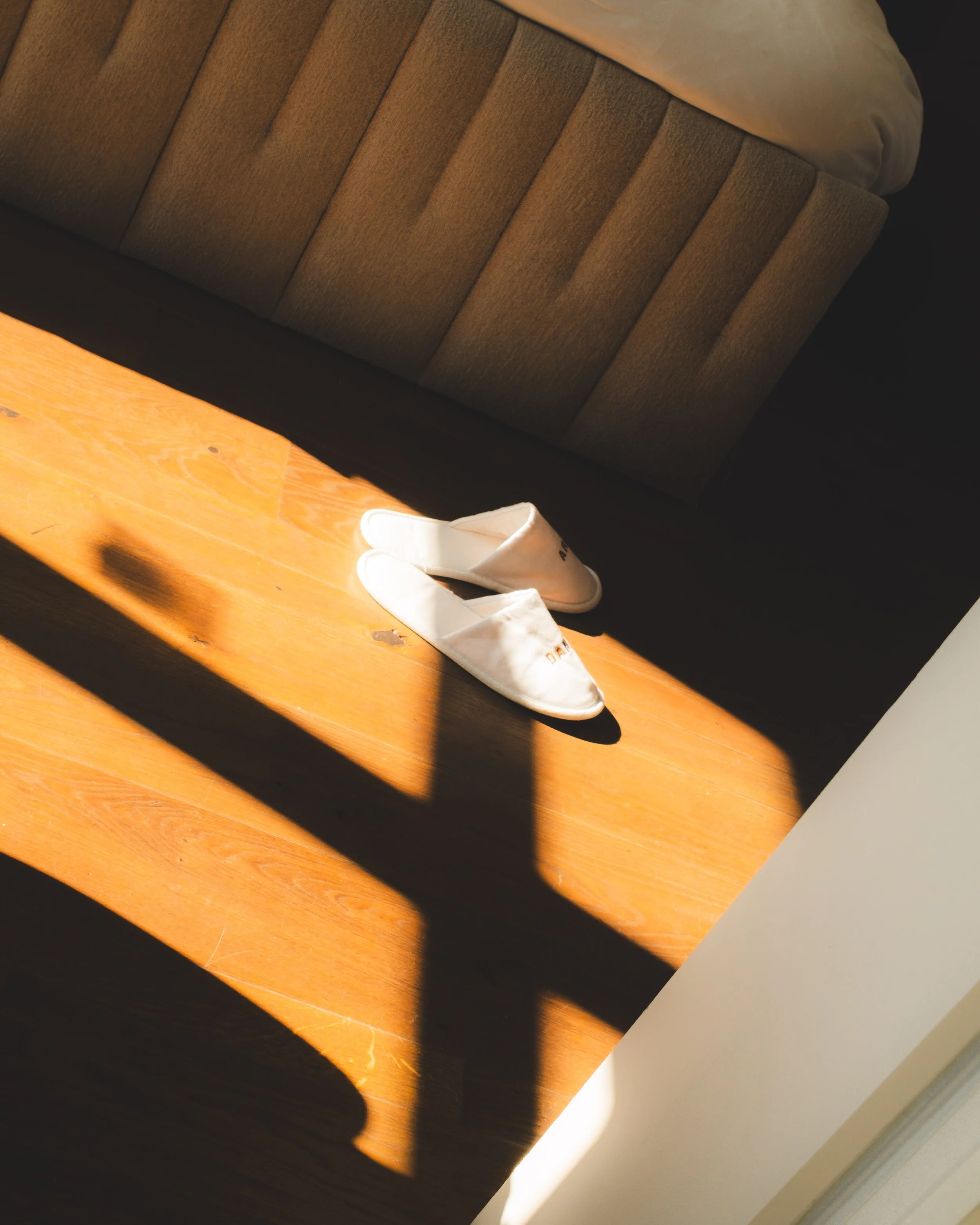 A pair of white fabric slip-on shoes on a wooden surface, with sunlight casting shadows.