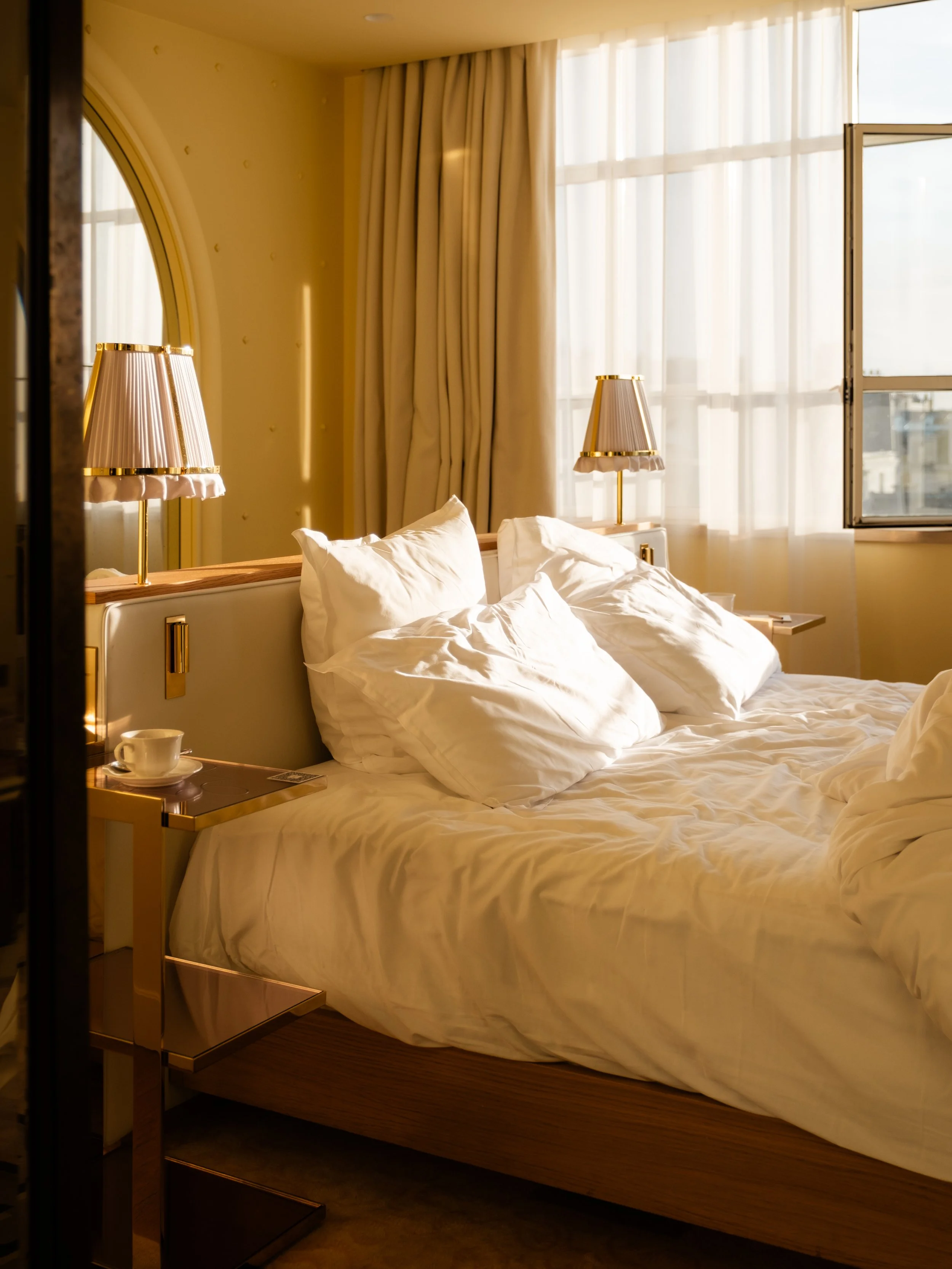 A neatly made hotel bed with white linens and pillows, illuminated by natural sunlight coming through open windows with sheer curtains, with bedside lamps and a cup on a small table. Lionel Chu - Hotel & Travel Photography