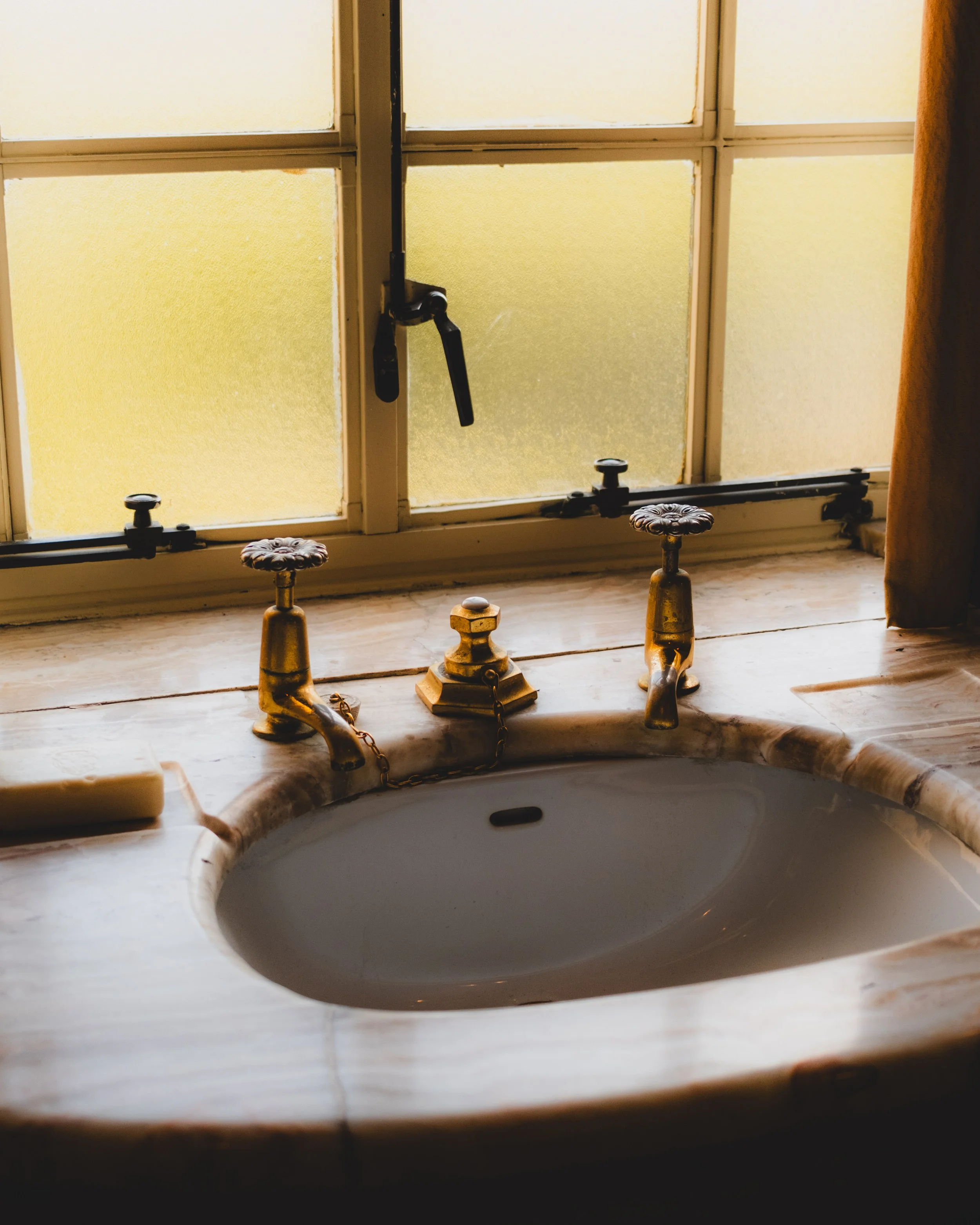 A bathroom sink with vintage brass fixtures, a marble countertop, and a window behind it. The window has closed frosted glass panes and a black handle, with a brown curtain partially visible on the right. Lionel Chu - Hotel & Travel Photographer