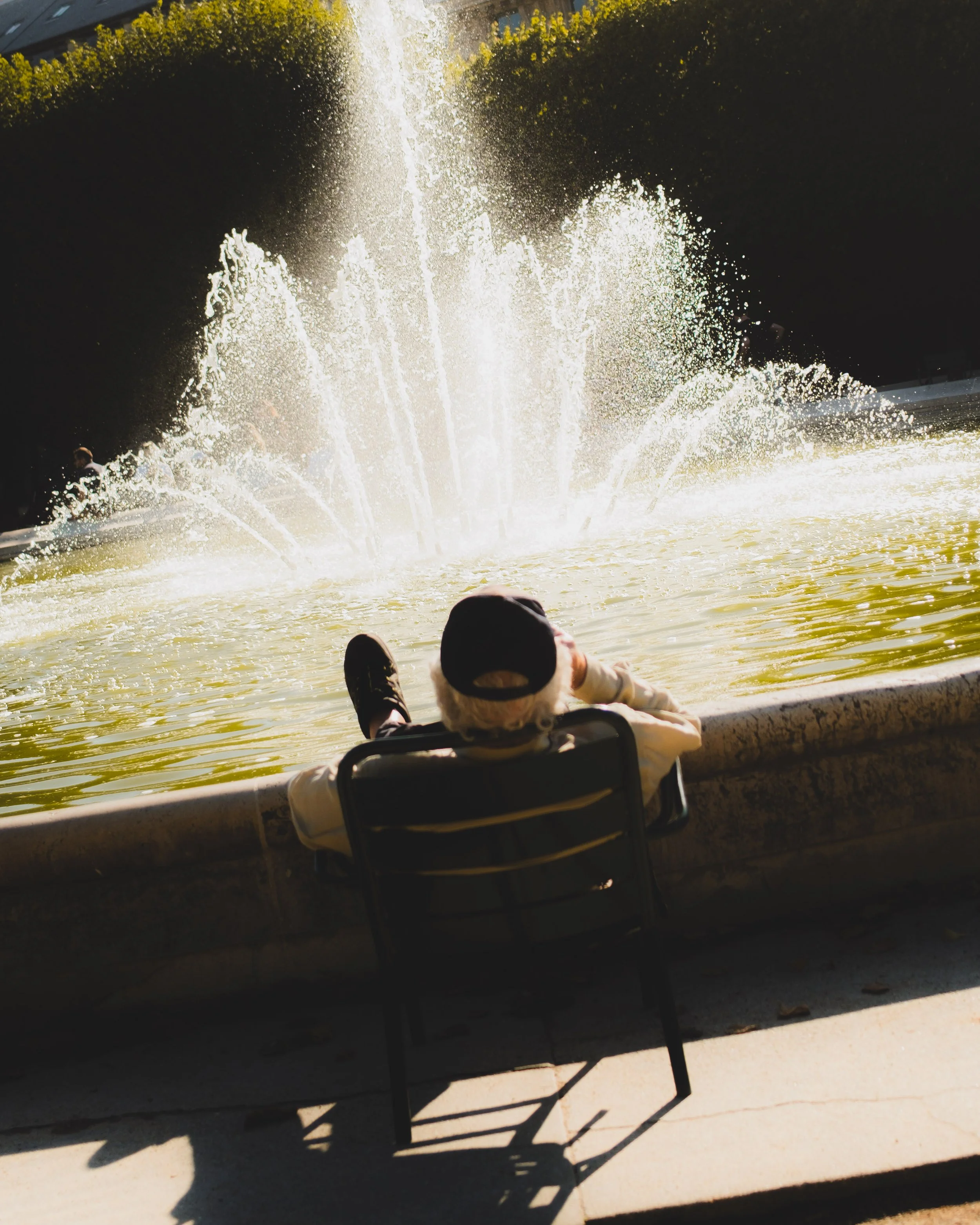 Old man with a cap sitting on a chair next to a fountain with water jets, outdoor park setting. Lionel Chu - Hotel & Travel Photographer.