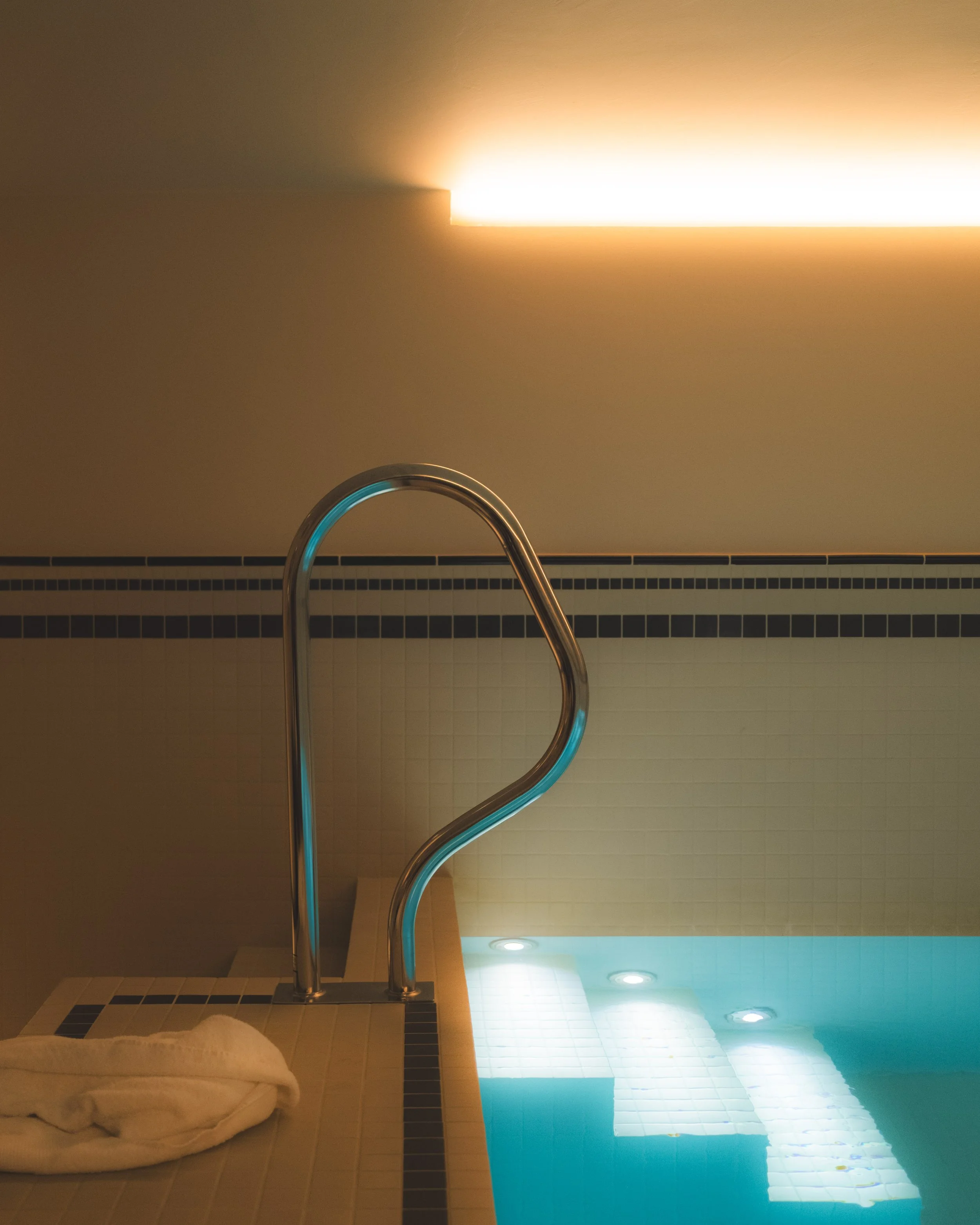 Indoor pool area with a metal handrail for swimming. The pool is illuminated with underwater lights, and there's a towel or blanket on the tiled floor beside the pool.