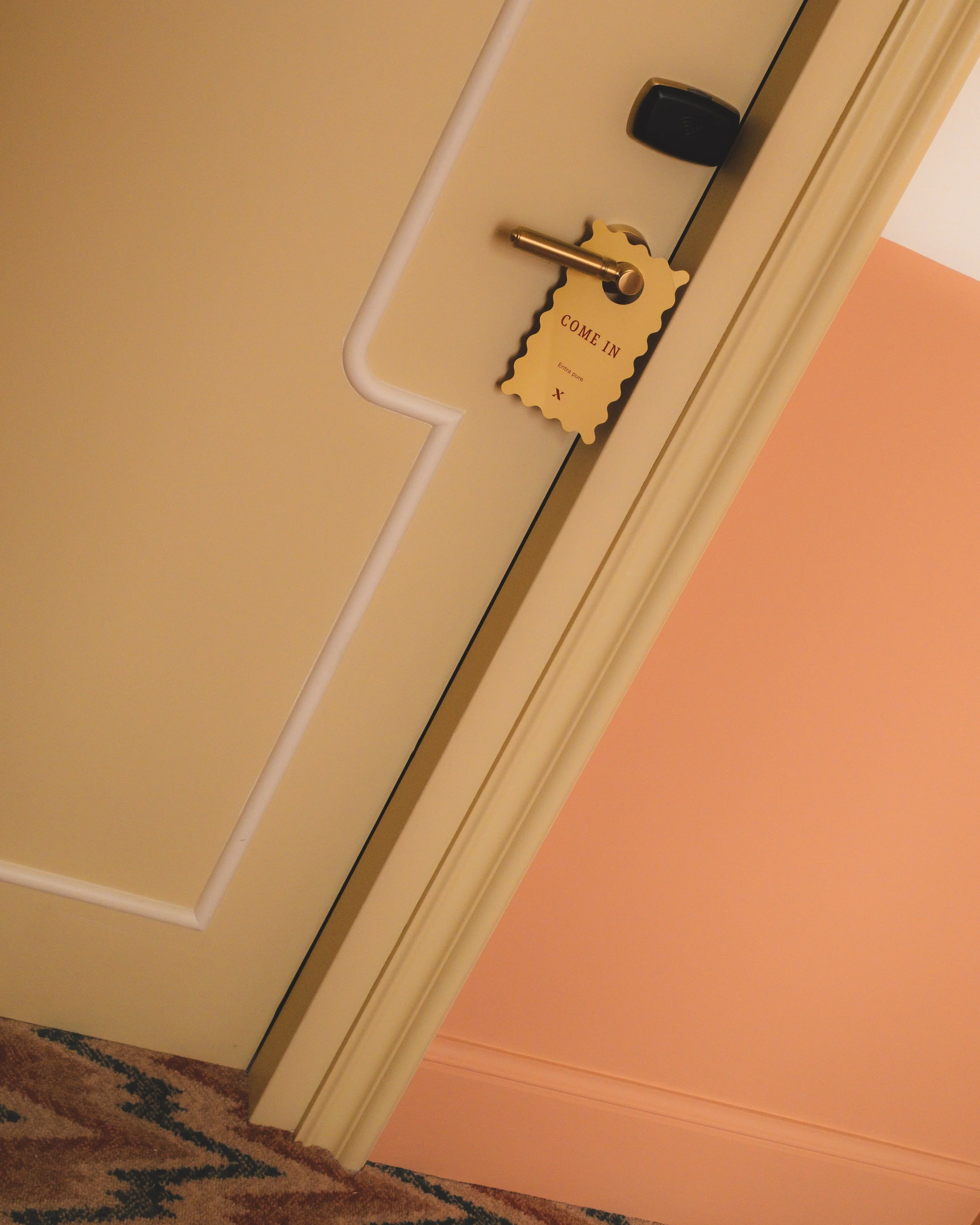 View looking up at a closed white door with a gold door handle and a black electronic lock. A yellow door hanger with scalloped edges hangs on the handle, reading "COME IN" and "Entre pure" with a small red cross at the bottom. part of a patterned carpet and peach-colored wall are visible at the bottom of the image.