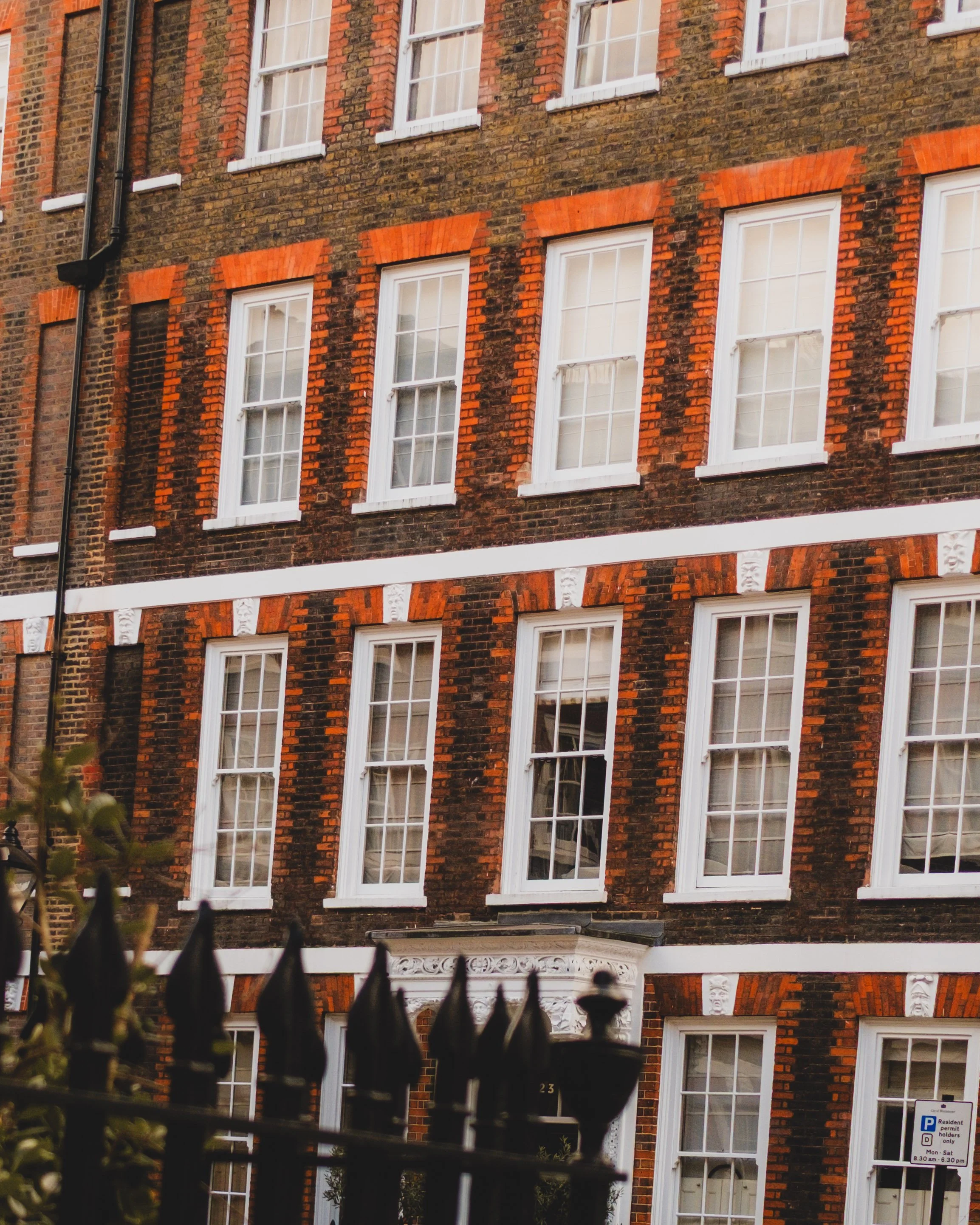 Three-story brick building with multiple white-framed sash windows, decorative white accents between floors, and a black iron fence at the front. The building has red brick arches above the windows and a black door with an ornate white decorative ped