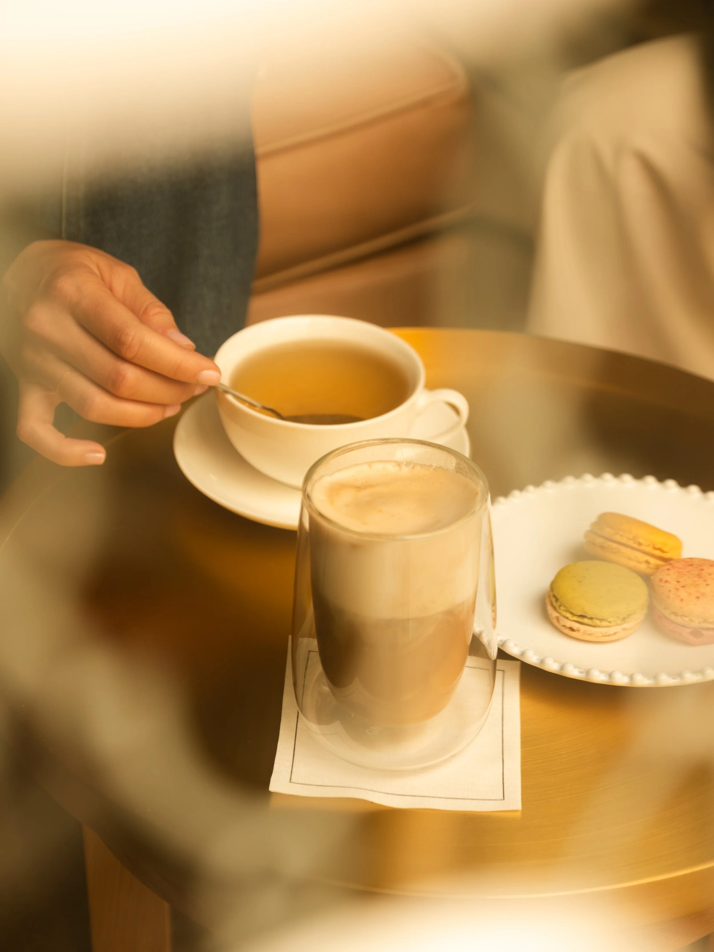 A person holding a spoon in a cup of tea, with a coffee mug, a glass of iced coffee, and a plate of assorted macarons on a wooden table. Lionel Chu - Hotel & Travel Photography