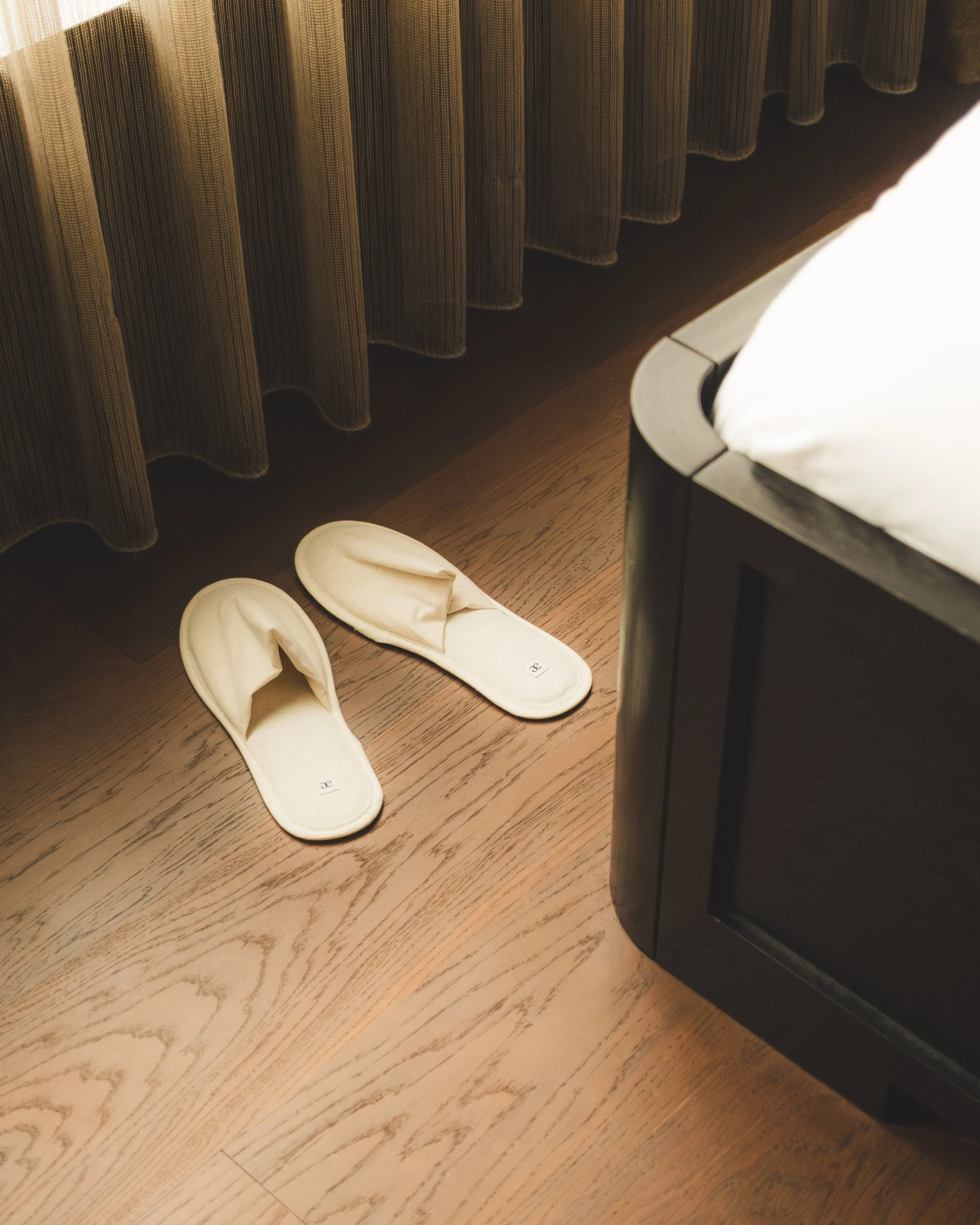 Pair of white slippers on a wooden floor next to a bed with a dark bedside table and a brown curtain.
