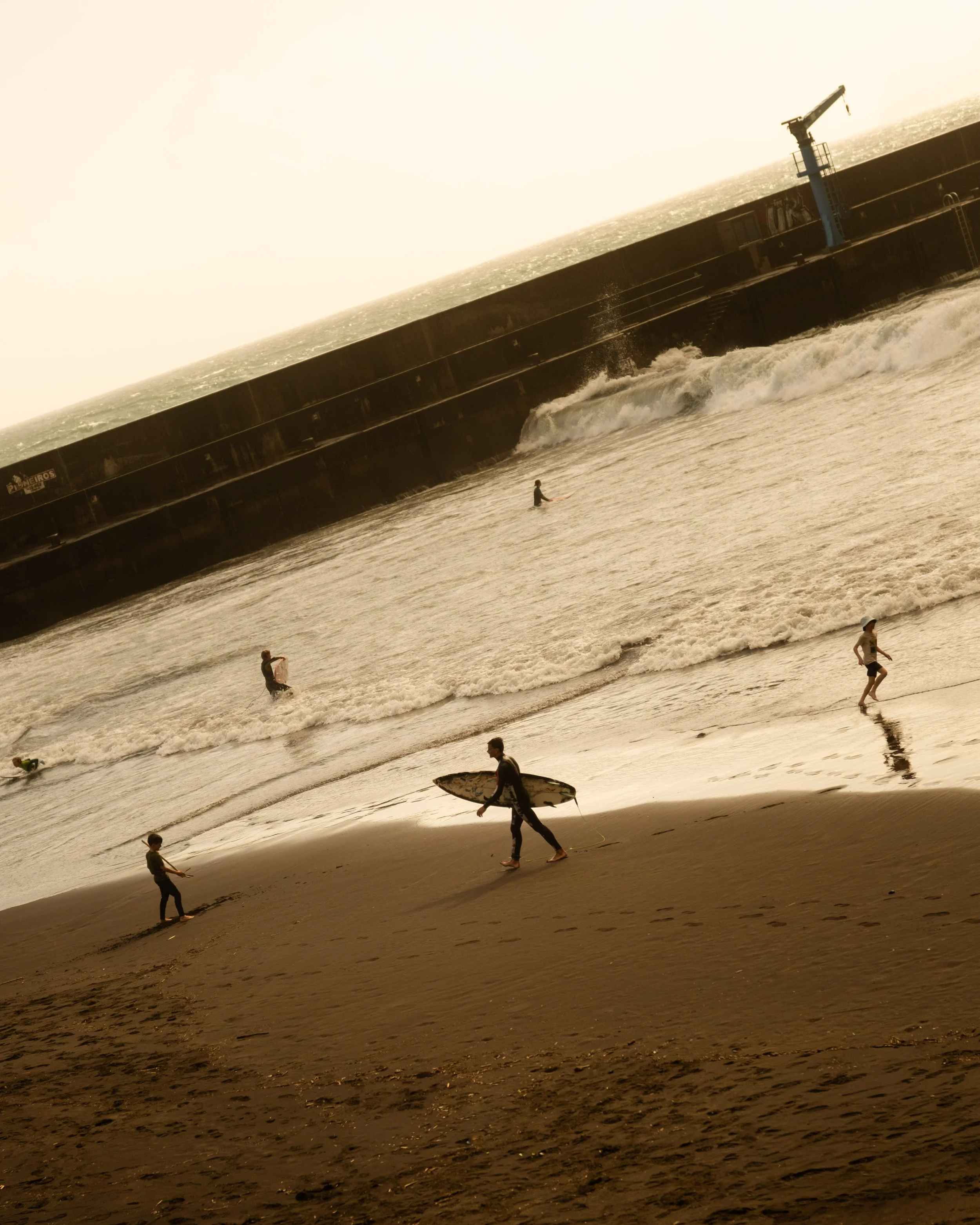 People at the beach, some surfing, some walking, with waves and a large ship or barge in the ocean in the background, and a hazy sky.