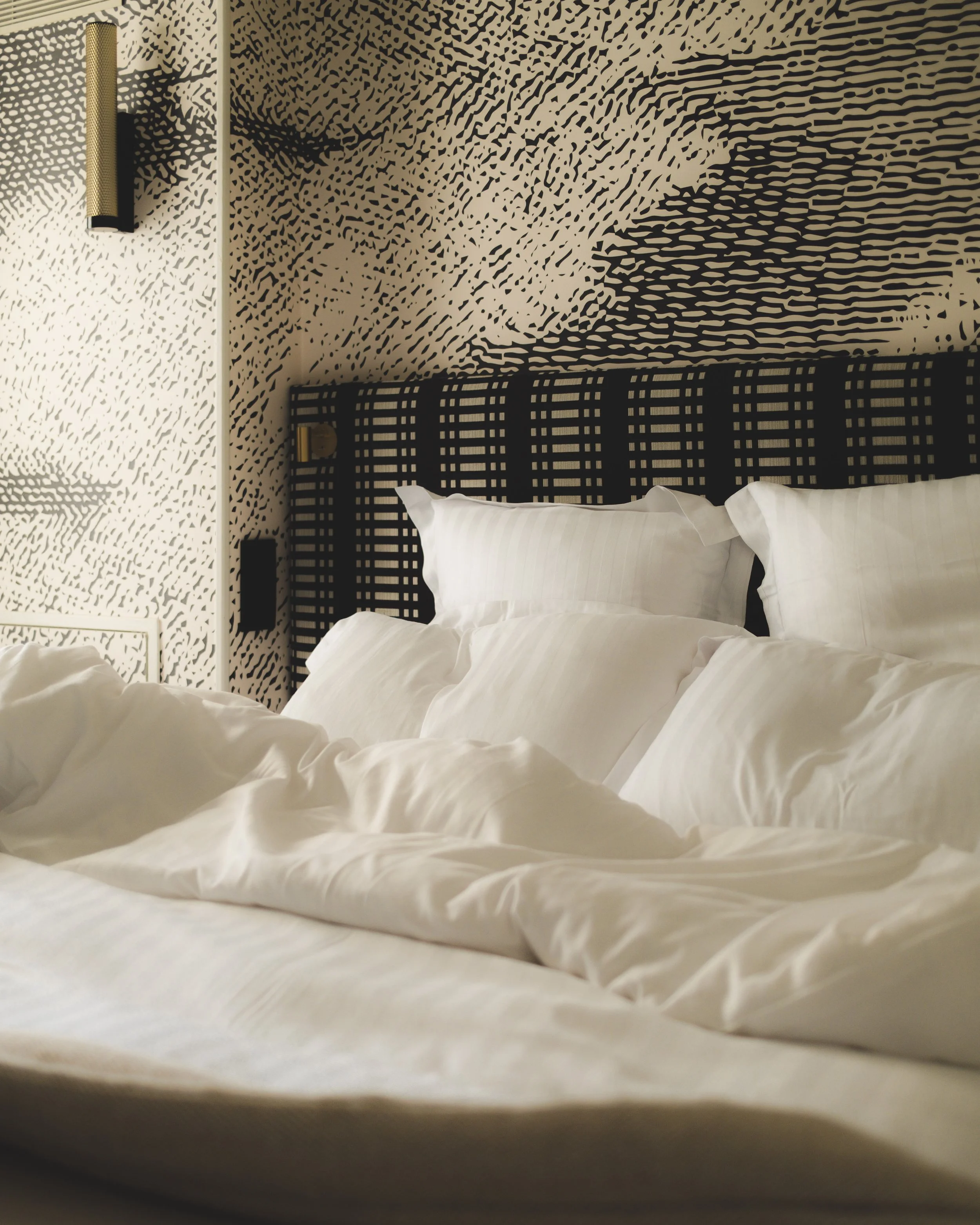 A view of a bed with white pillows and white bed sheets, a black headboard, and a textured wall with a black and beige abstract pattern. There is a gold cylindrical wall-mounted light fixture on the left side of the image.