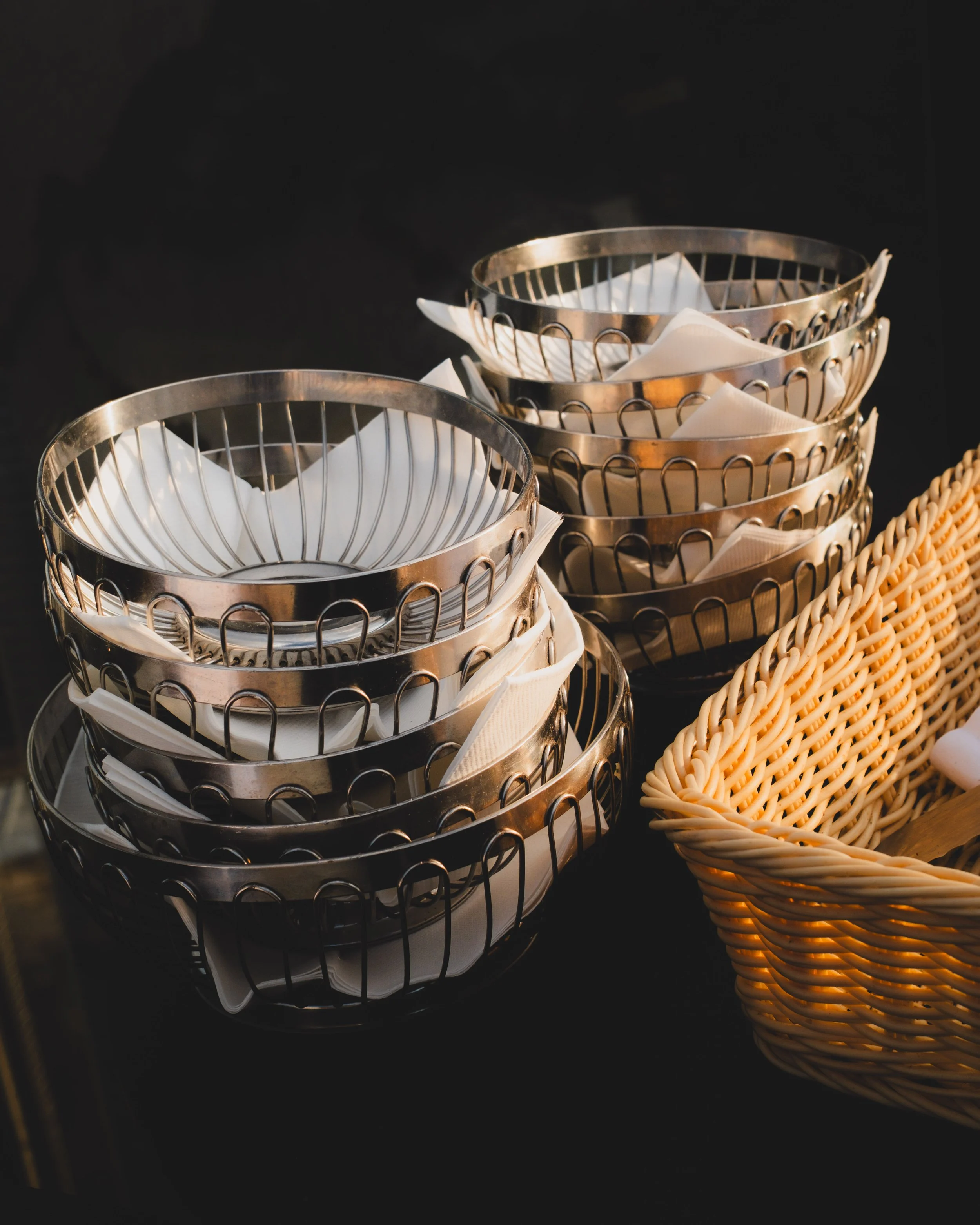 Stacked metal baskets with white napkins inside, adjacent to a woven basket, on a black surface.