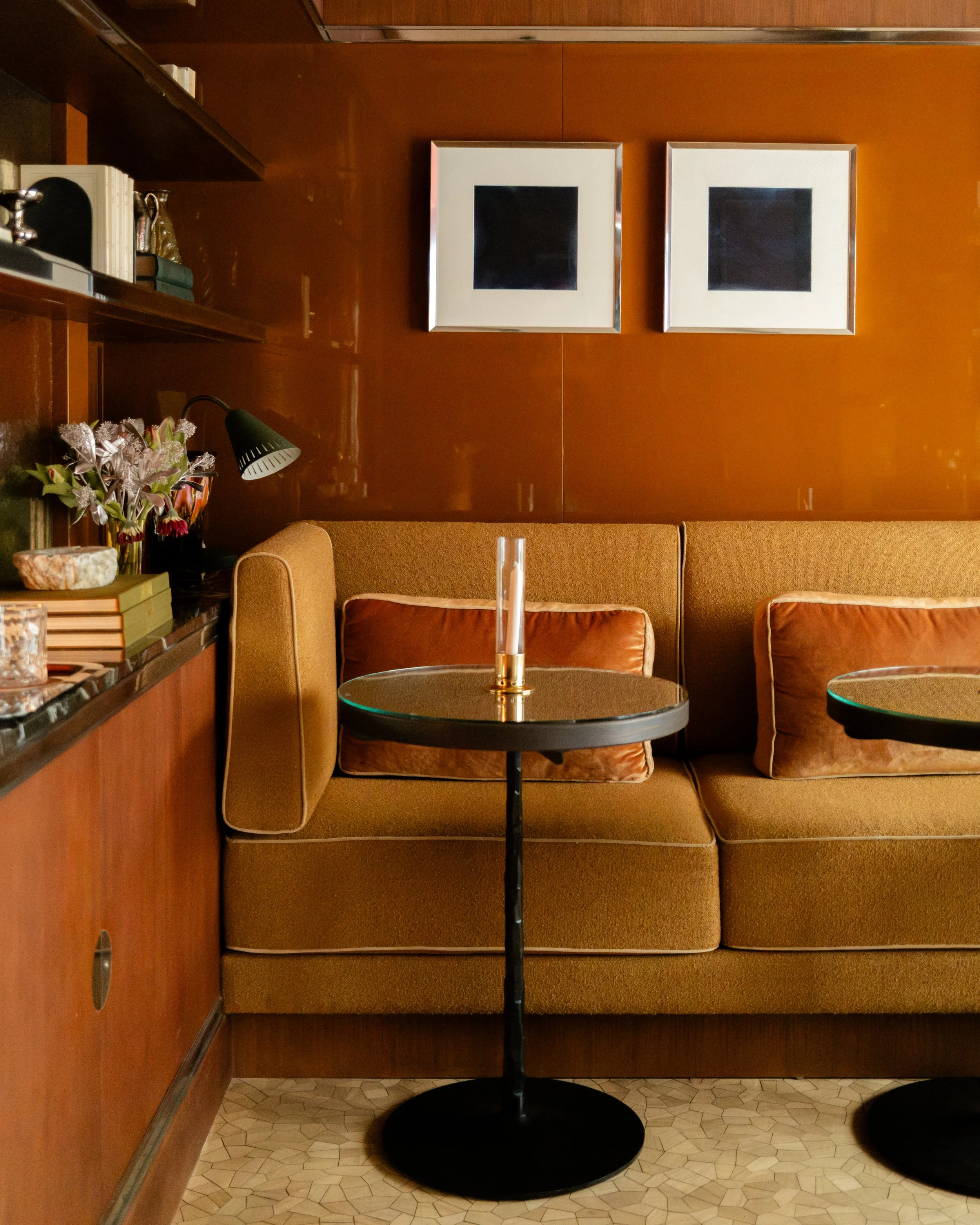 A cozy indoor seating area with a brown cushioned bench, pillows, and a glass-topped table with a candle holder. On the left, shelves hold books, flowers, and decorative items. Two framed black squares hang on the wooden wall behind. Lionel Chu