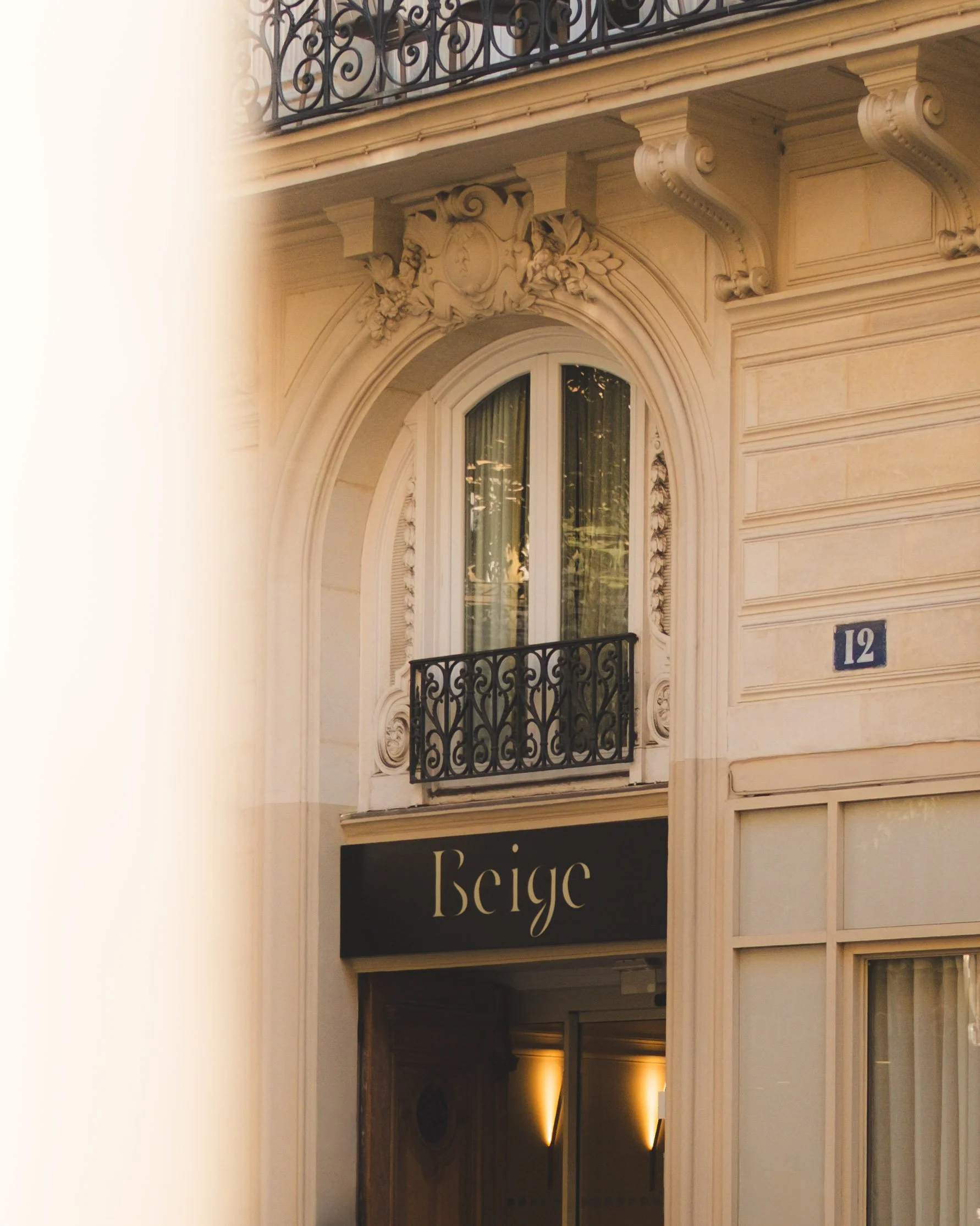 The image shows the upper part of a building facade with ornate architectural details, a small corner balcony with a wrought iron railing, a window with curtains, and a sign that reads 'Beige'. The building features intricate carvings and mouldings around the window and balcony, and the number 12 is displayed on the wall.