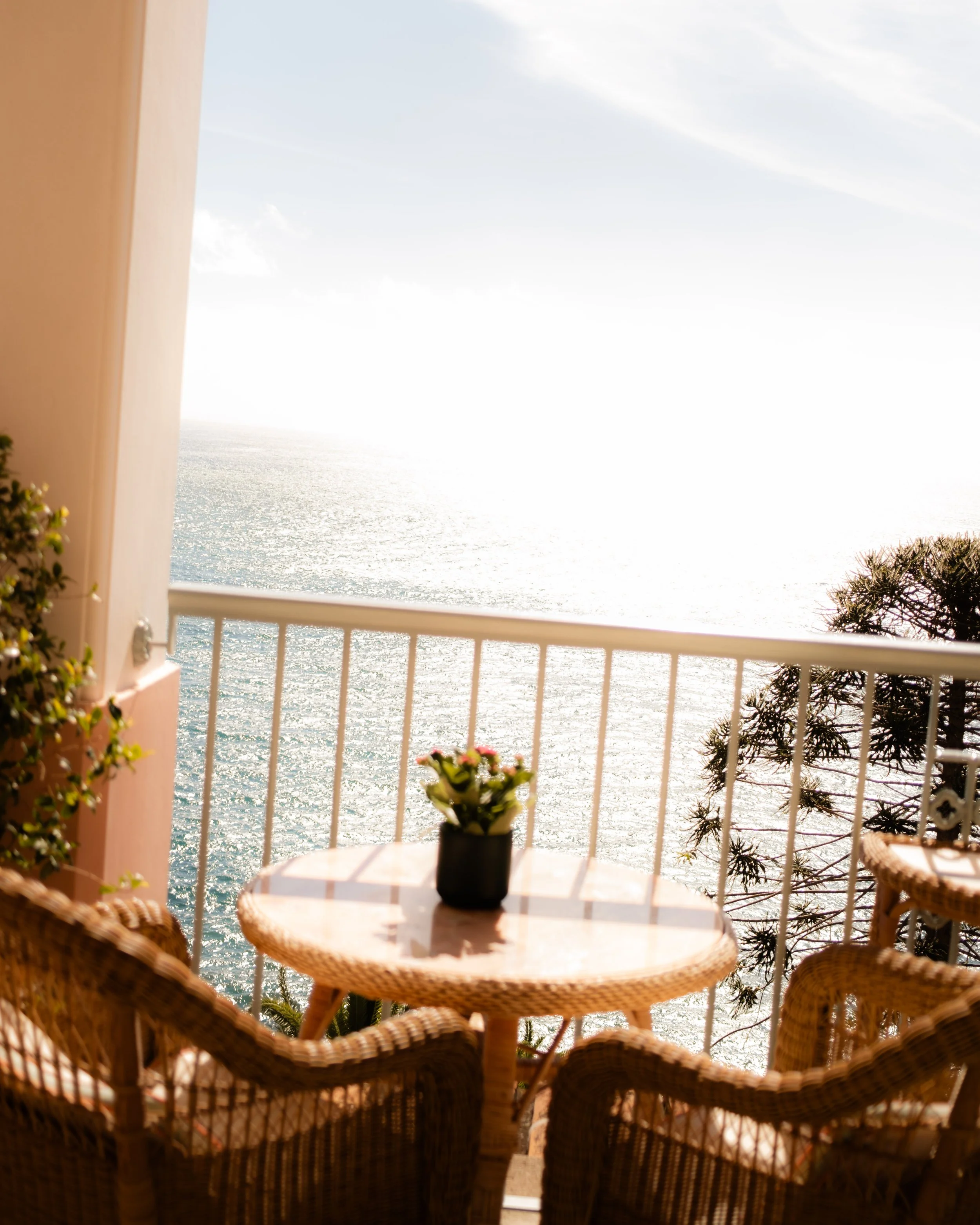 View from a balcony overlooking a shimmering ocean with a partly cloudy sky. The balcony has wicker chairs and a small round table with a potted plant in the center. The scene is bright and sunny, suggesting a warm day.