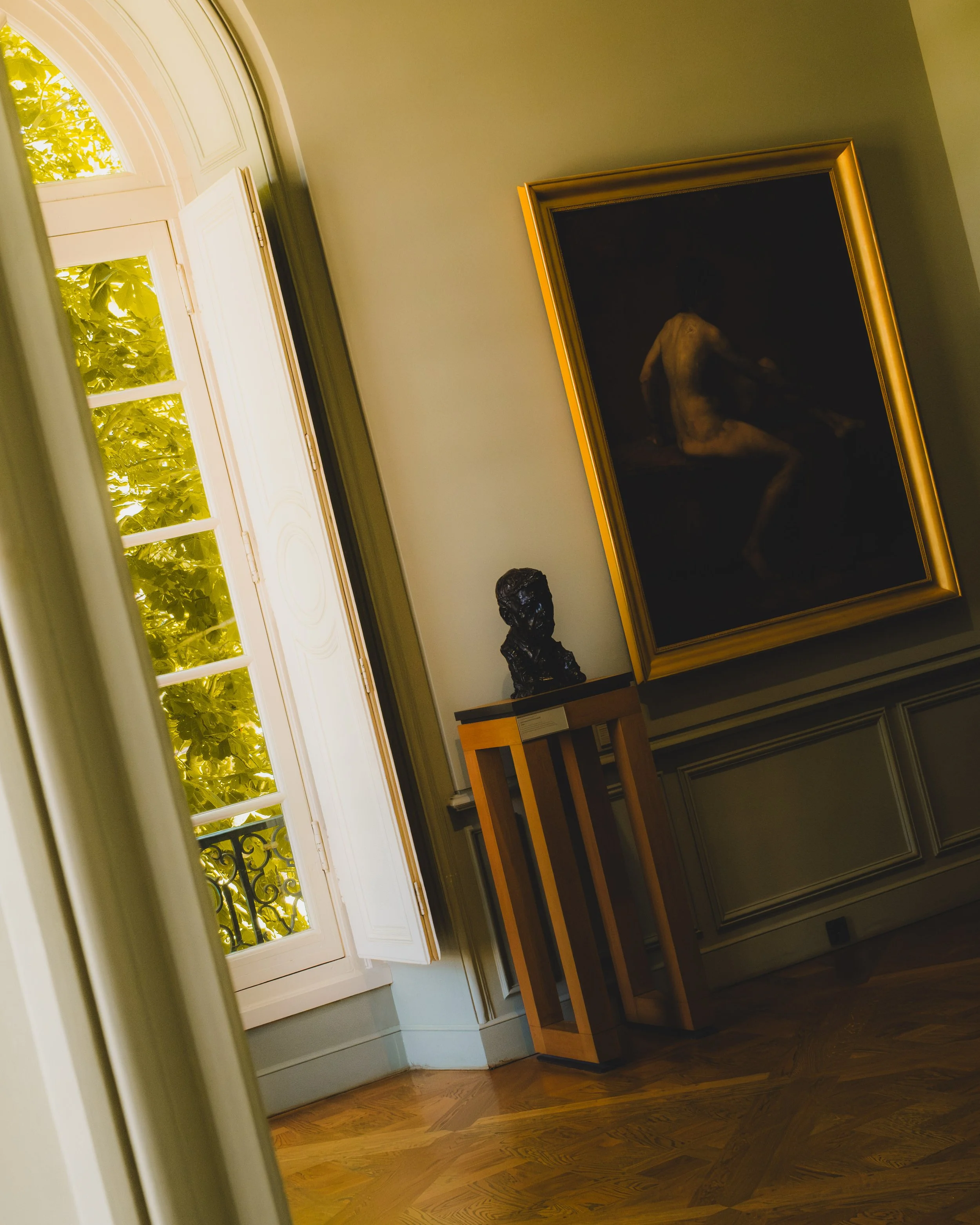 Interior of a room with an open arched window, a framed painting of a nude figure, and a bust sculpture on a wooden pedestal. Lionel Chu - Hotel & Travel Photographer.