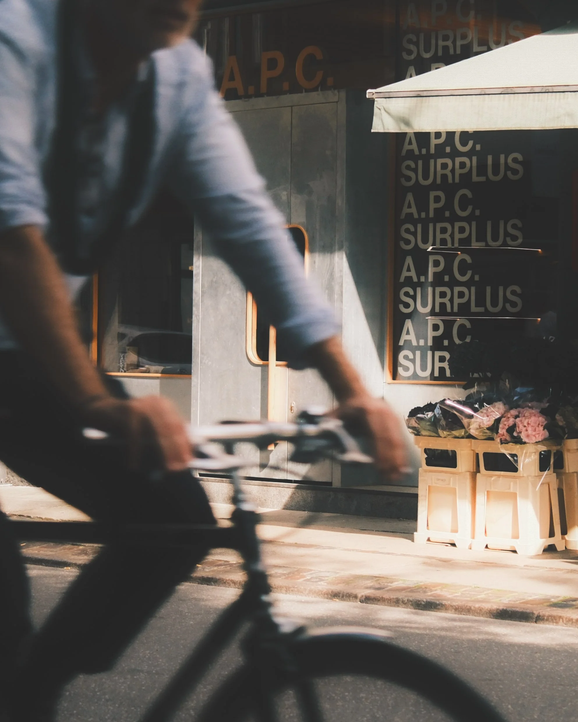A person riding a bicycle past an APC clothing store with some crates with flowers outside.