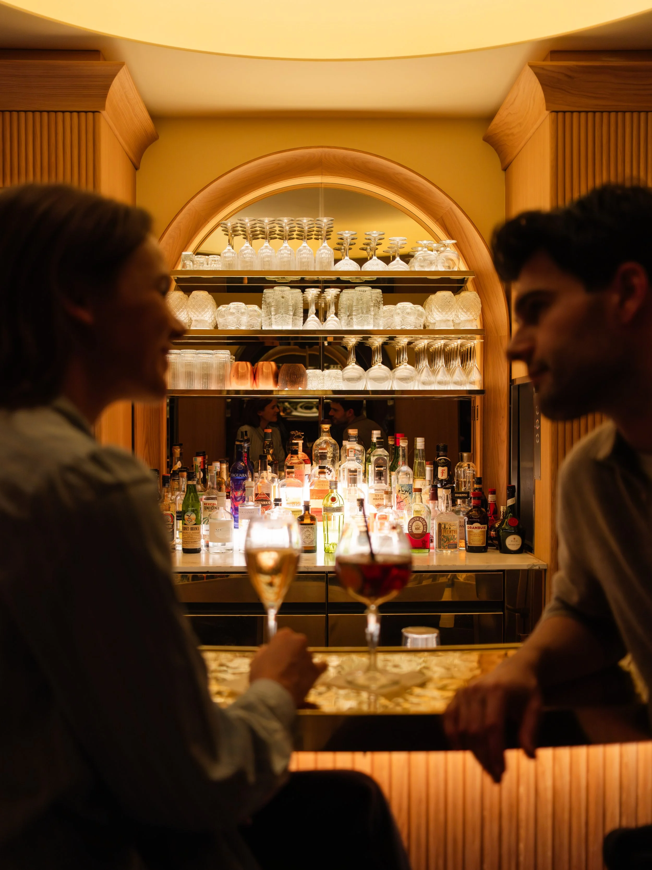 Two people are sitting at an hotel bar with drinks, in a warmly lit bar or restaurant with wooden decor and glass shelves filled with glasses and bottles. Lionel Chu - Hotel & Travel Photography