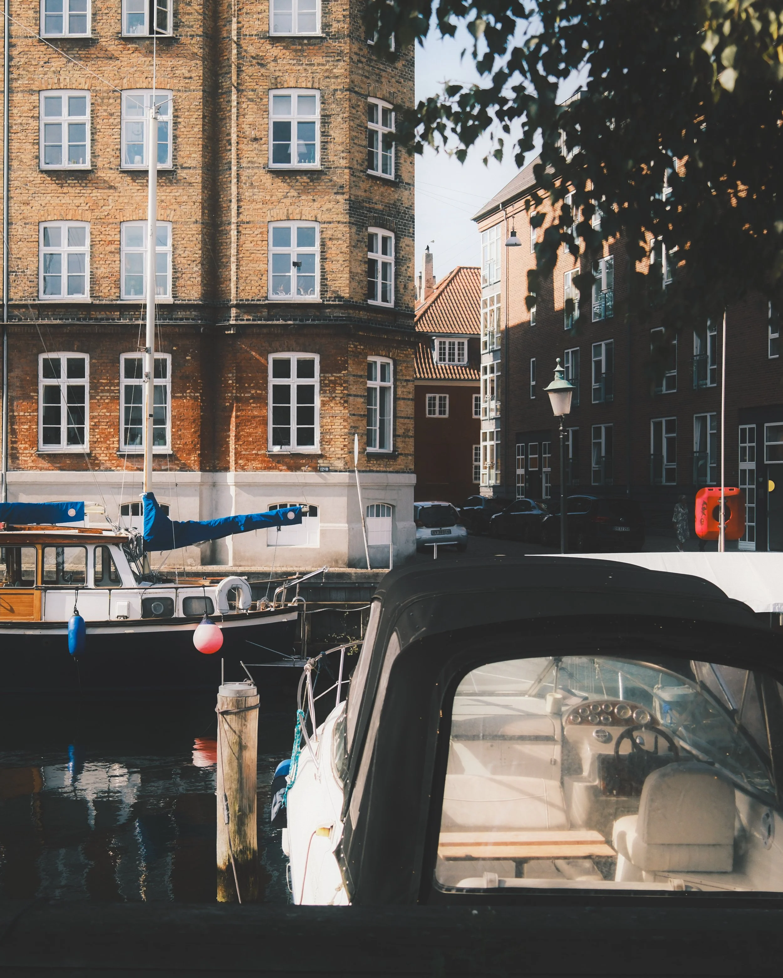 View through the window of a boat docked at a marina showing several brick buildings with white framed windows. There are boats moored in the water, some with blue covers, and a streetlamp is visible. The scene suggests a peaceful harbor in an urban 